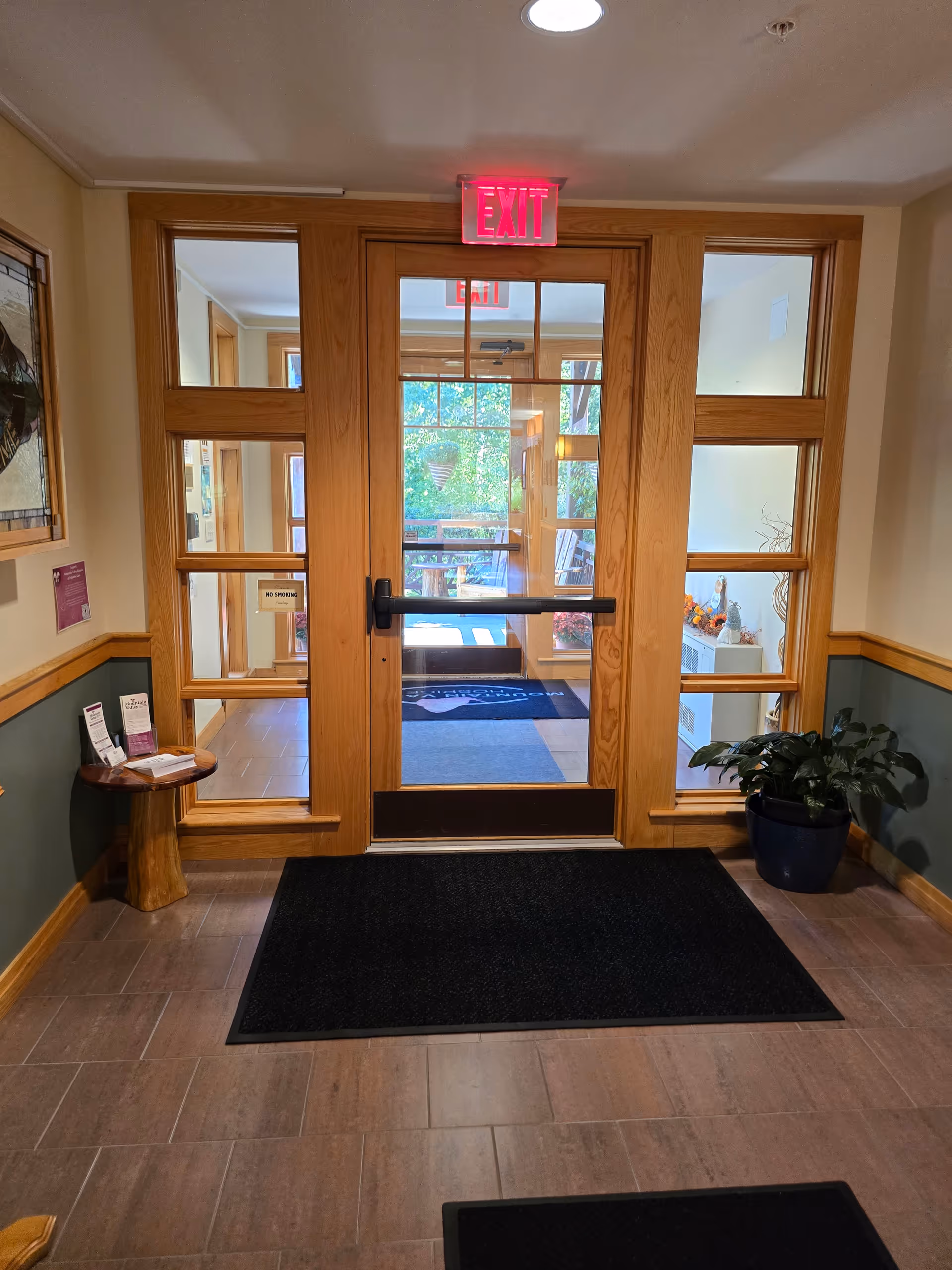 Wooden double doors with glass panes, exit sign, and a black mat in a lobby entry.