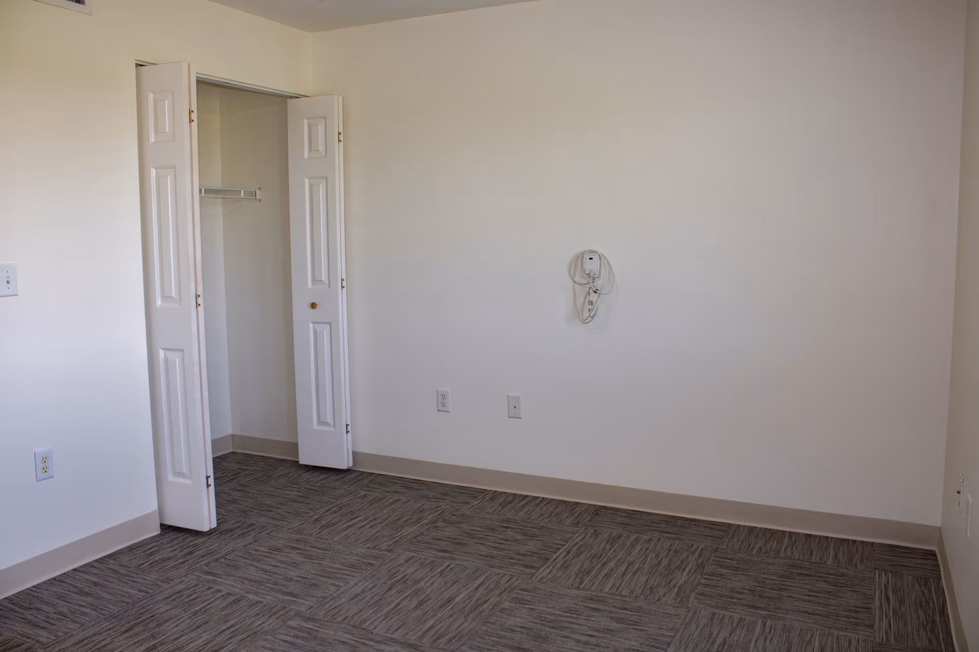 Empty bedroom with closet doors open, beige carpet, and plain white walls.