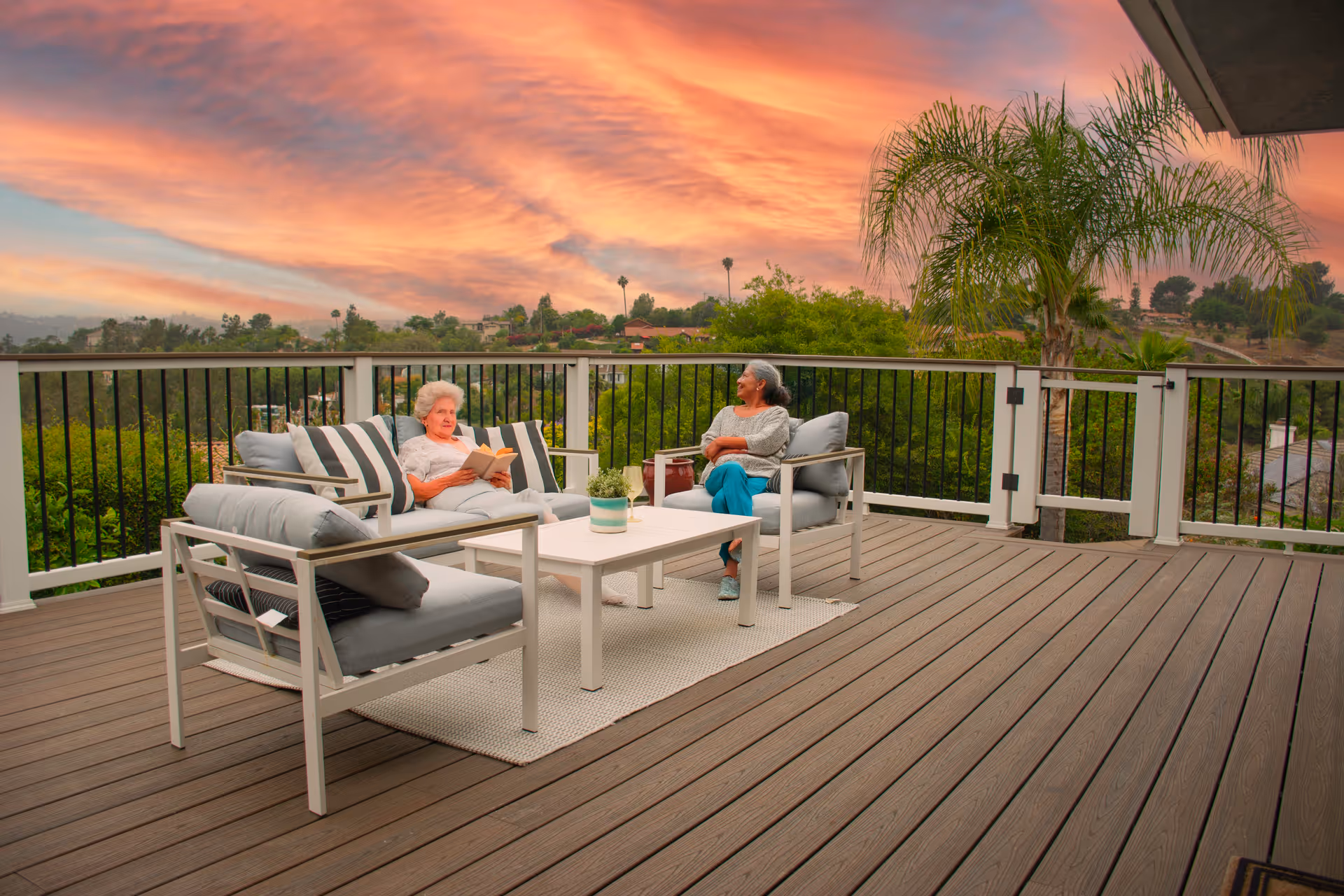 Two elderly women relax on outdoor lounge furniture on a deck at sunset.