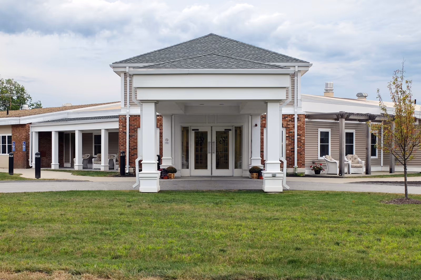 Front exterior view of Holden Rehabilitation & Skilled Nursing Center showing a covered entrance with white pillars, brick and siding walls, and a grassy area in front under a cloudy sky.