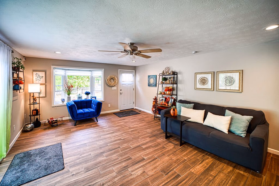 A bright and spacious living room with wood flooring, a ceiling fan, a blue armchair near a large window, a black sofa with white and light blue pillows, a small black side table with a pumpkin decoration, and a black shelving unit with various decorative items. The walls are light-colored with framed artwork and a wreath near the white front door.