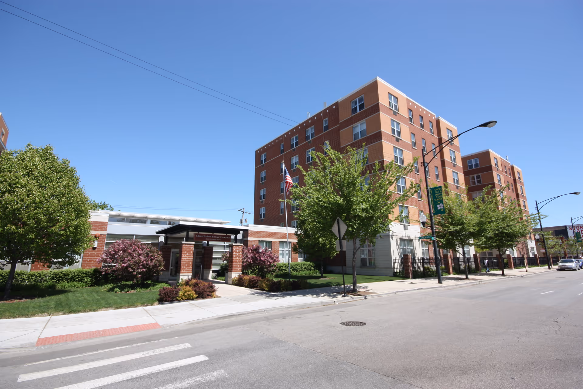 Street view of a multi-story brick residential building with an entrance canopy, trees, and an American flag under a clear blue sky.