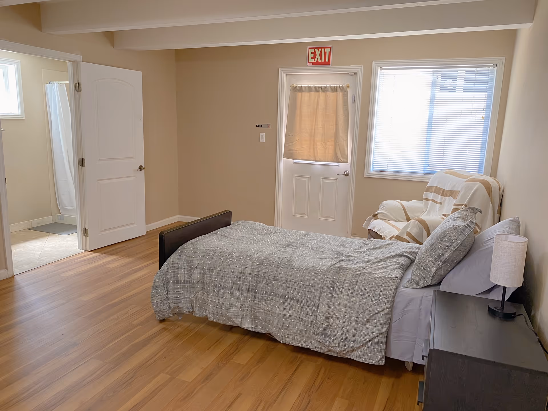 Bedroom with a bed, nightstand, chair, window, and doorway in a home care facility.