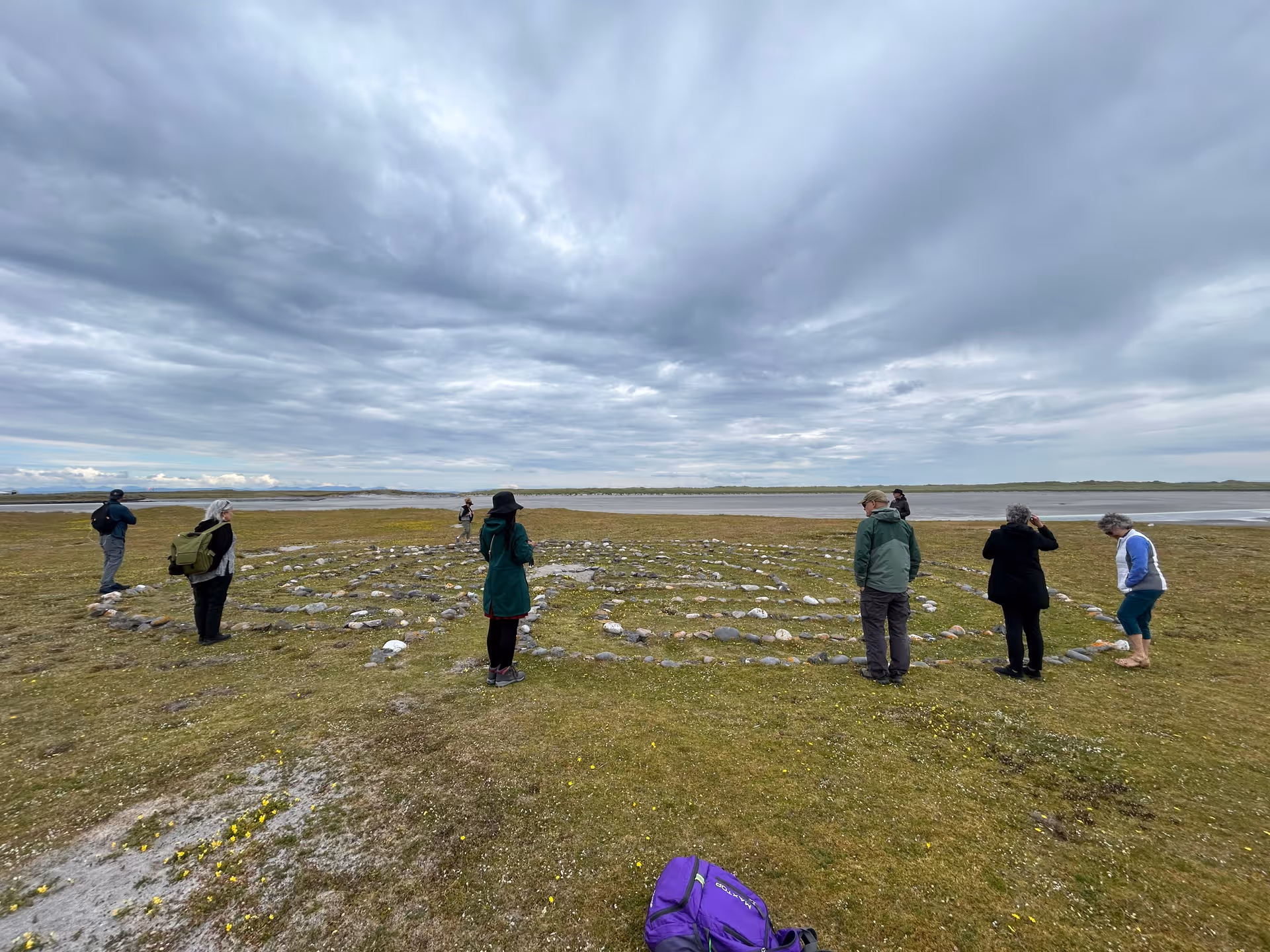 Group of people outdoors near a shoreline, standing around a circular stone arrangement.