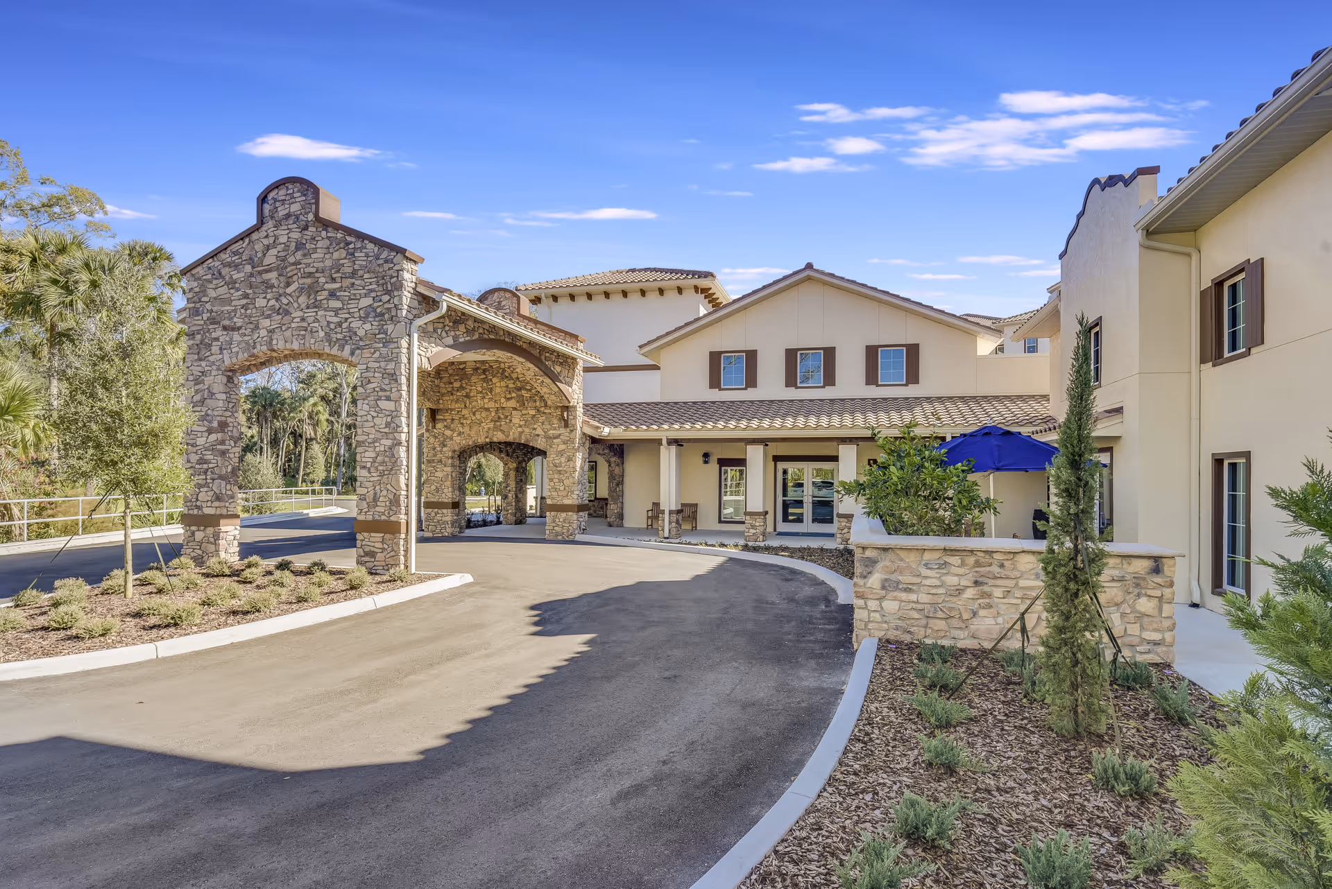 Exterior view of Tuscan Gardens of Palm Coast Senior Living facility showing a stone archway entrance, beige building with brown shutters, a curved driveway, landscaped plants, and a blue umbrella on a patio area under a clear blue sky.