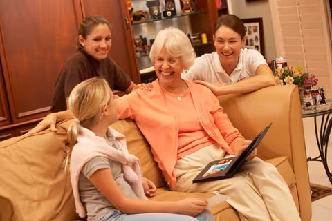Elderly woman on a beige sofa with a laptop, surrounded by smiling caregivers.