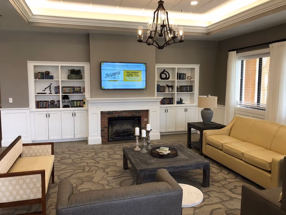 A cozy living room with a beige sofa, two gray armchairs, and a wooden bench with patterned cushions. A wooden coffee table with candles and a tray sits in the center on a patterned carpet. The room features a white fireplace with a mounted flat-screen TV above it, flanked by built-in white bookshelves filled with books and decorative items. A window with white curtains allows natural light to enter, and a chandelier hangs from the ceiling.