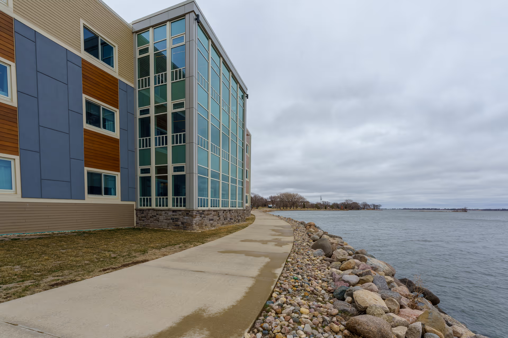 Modern multi-story senior living building along a paved path beside a rocky lakeshore.
