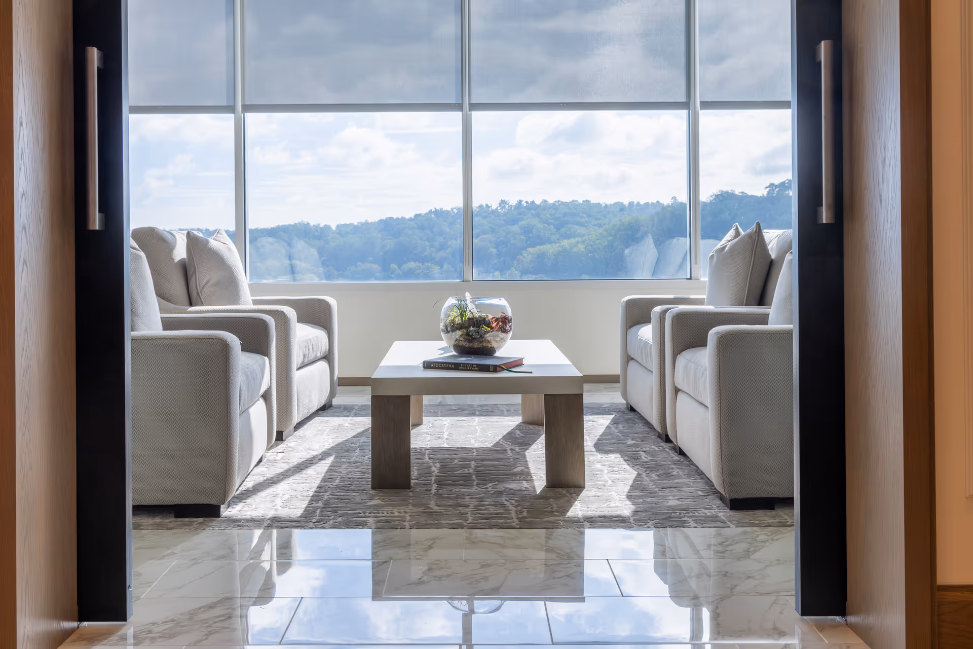 Two light armchairs flank a coffee table beside a large window overlooking hills.