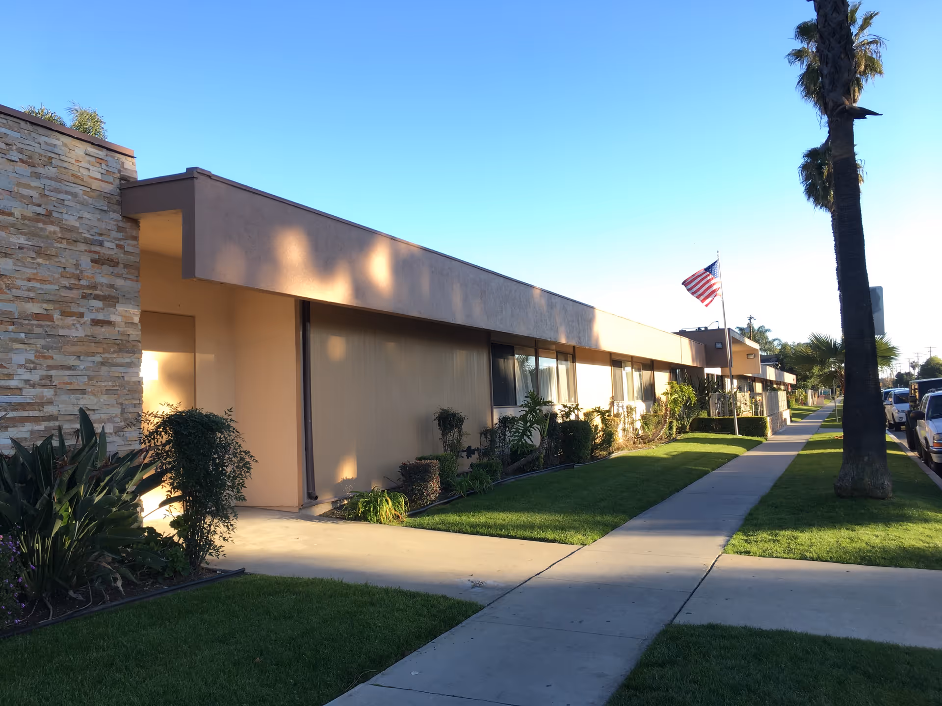 Single-story senior living facility exterior with beige walls, sidewalk, landscaping, and American flag.