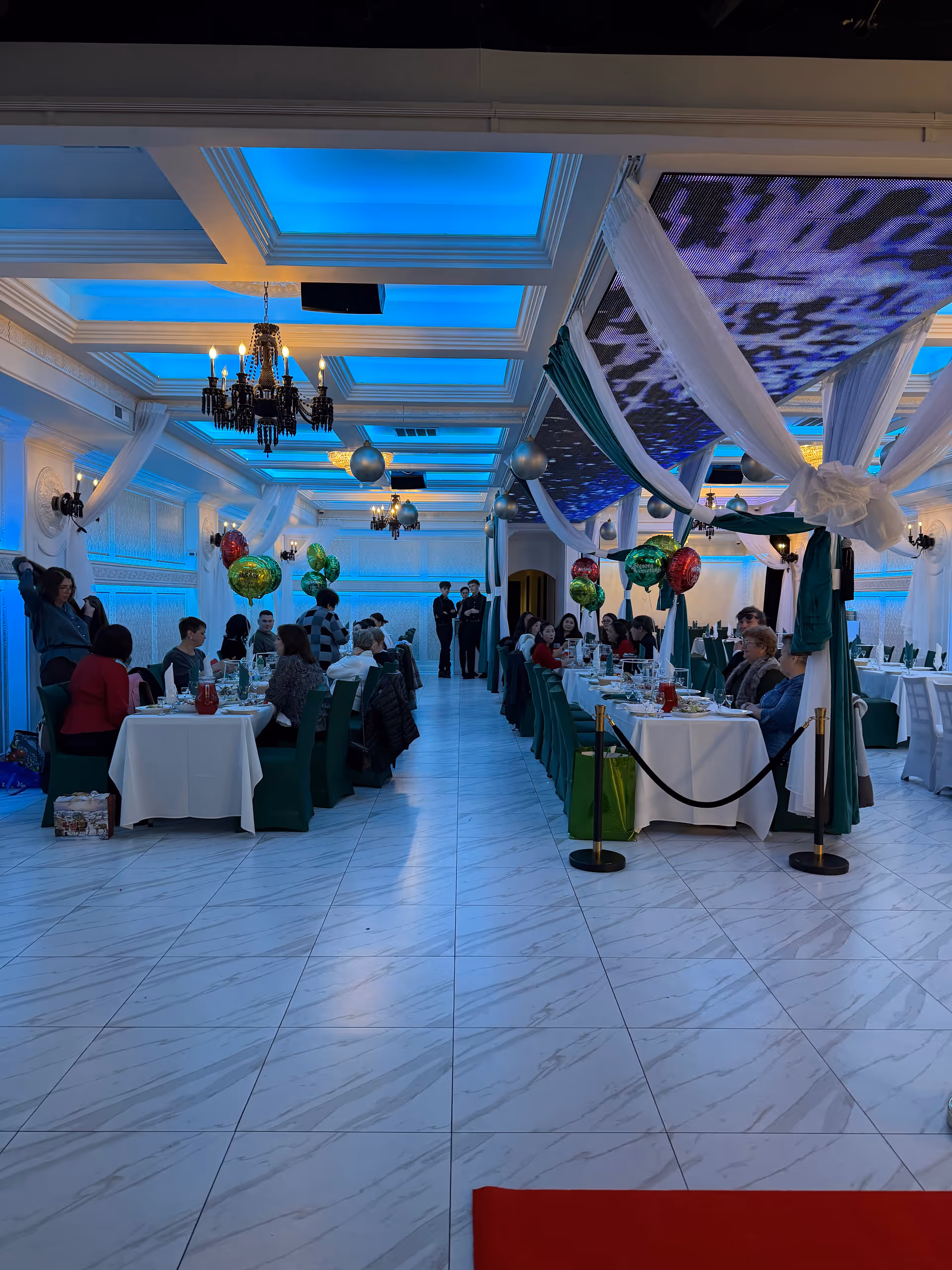 A large, well-lit dining hall with decorated tables, balloons, and guests dining together.