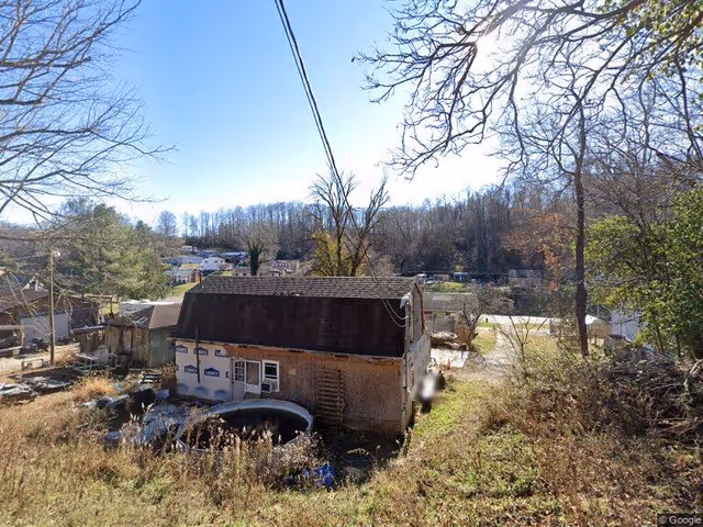 A small weathered outbuilding and overgrown yard on a hillside with trees and houses in the background under a clear sky.
