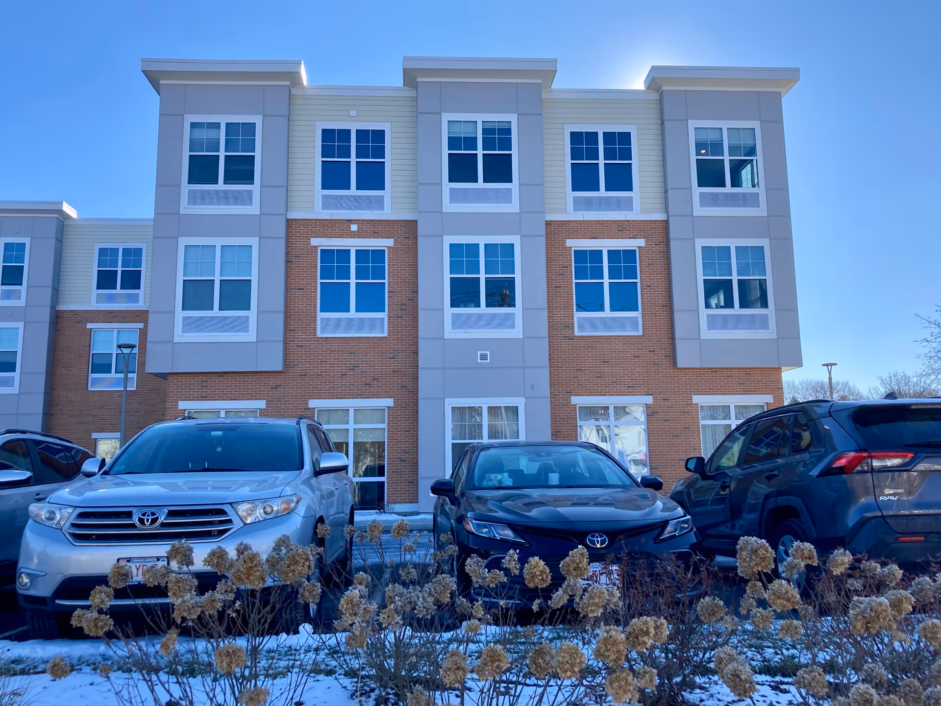 Three-story brick-and-siding residence building facade with parked cars and light snow in front.
