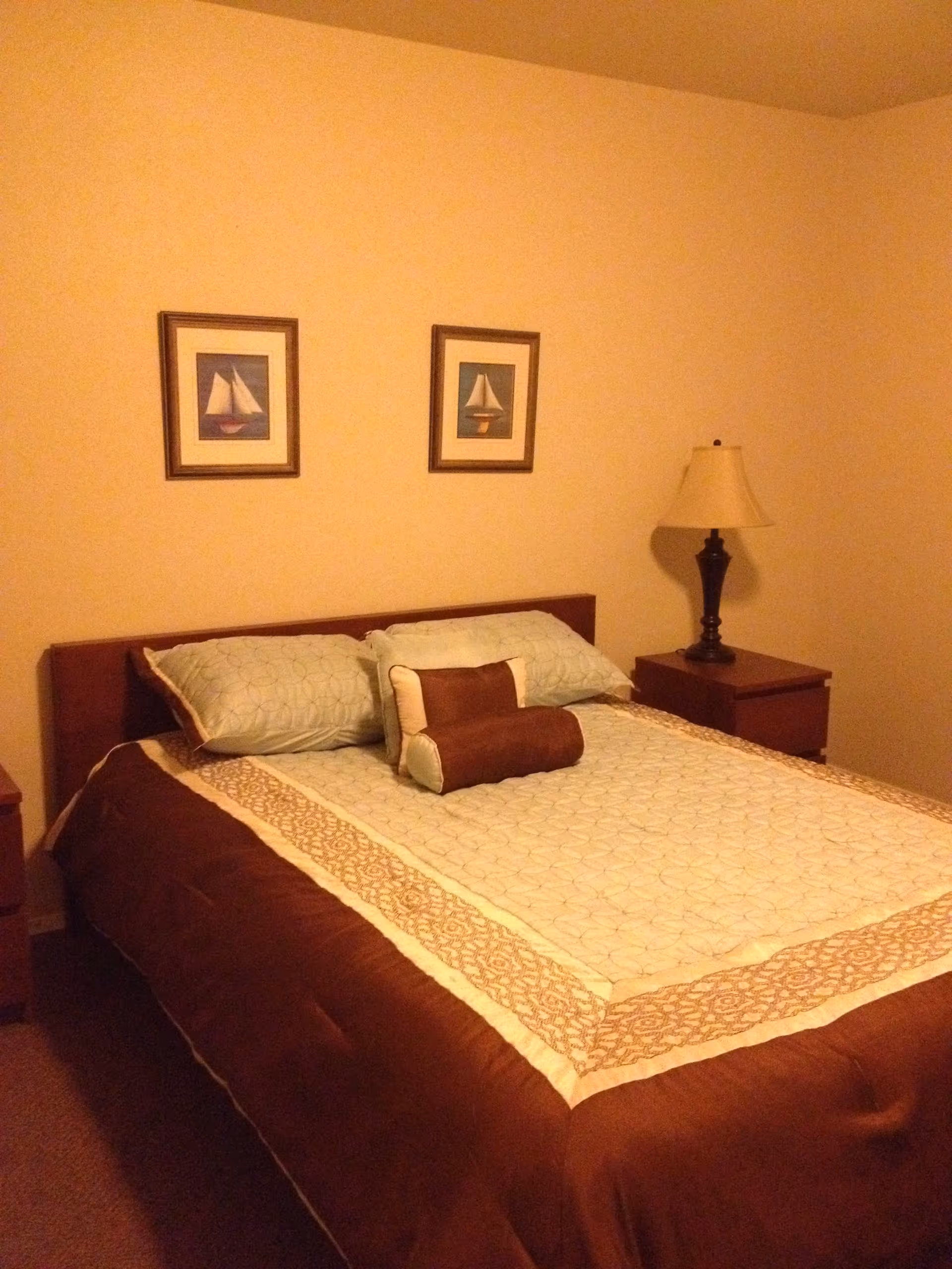 A bedroom with a neatly made bed featuring a brown and beige patterned comforter, two light blue pillows, and two decorative brown and beige pillows. There are two framed pictures of sailboats hanging on the beige wall above the bed. A wooden nightstand with a lamp is positioned to the right of the bed.
