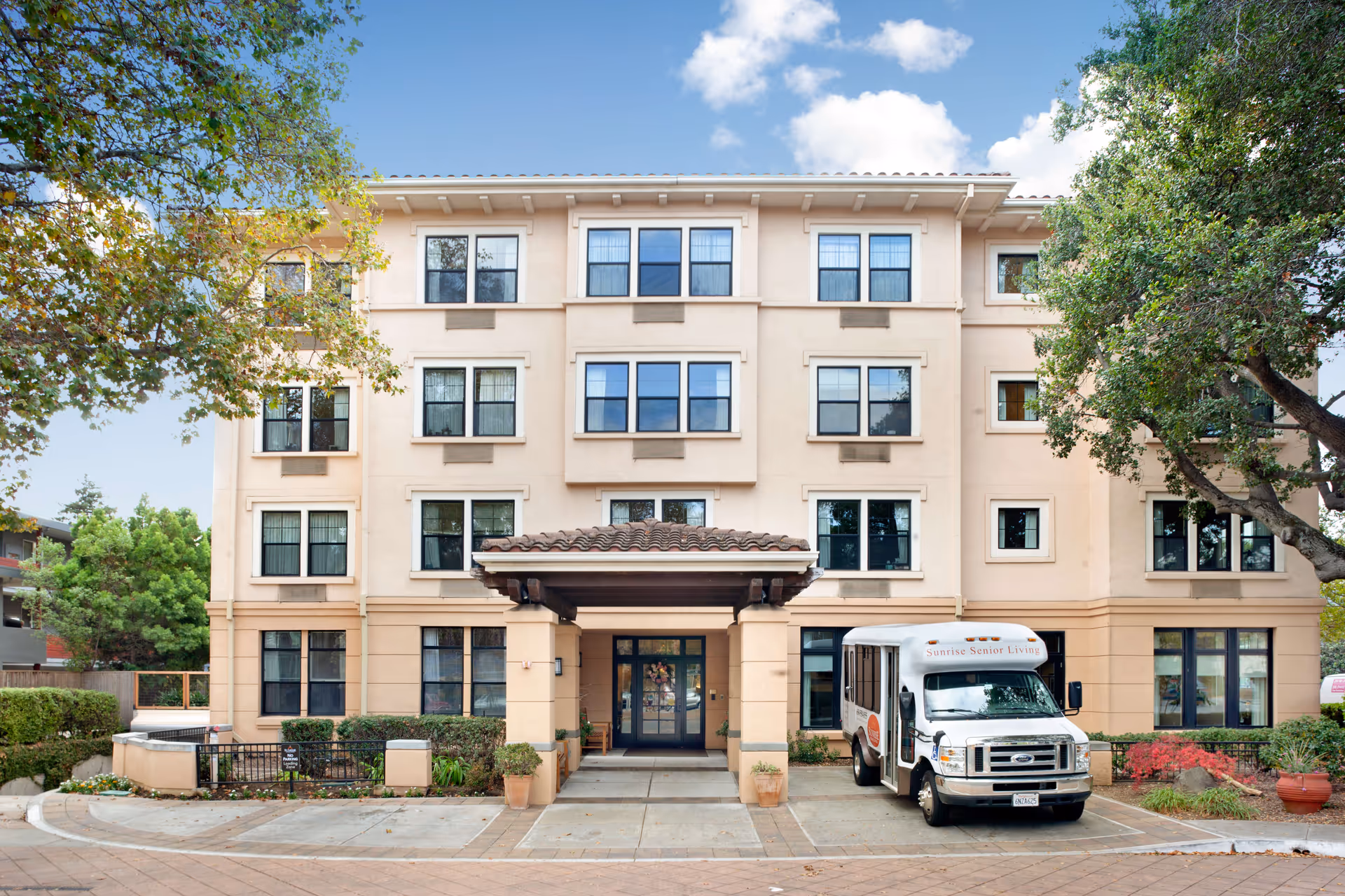 Three-story beige Sunrise Senior Living building with a driveway, entrance, and parked shuttle bus.