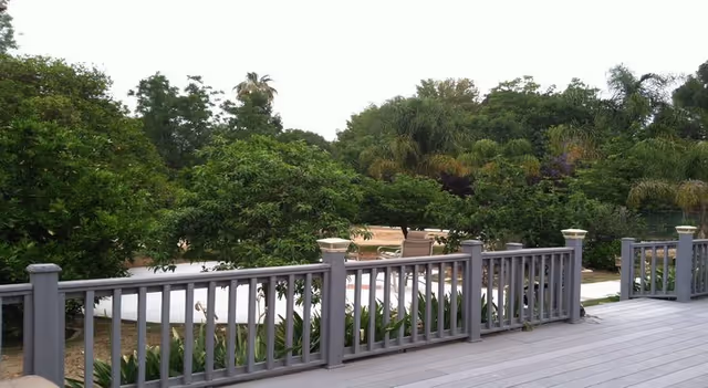 Wooden deck railing overlooks a landscaped outdoor area with lounge chairs and trees.