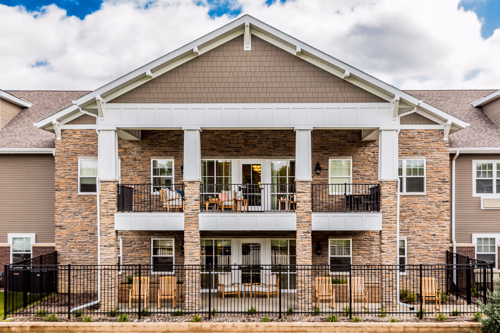 Exterior view of a senior living facility building with stone and siding facade, featuring two levels of balconies and patios with outdoor seating, under a partly cloudy sky.