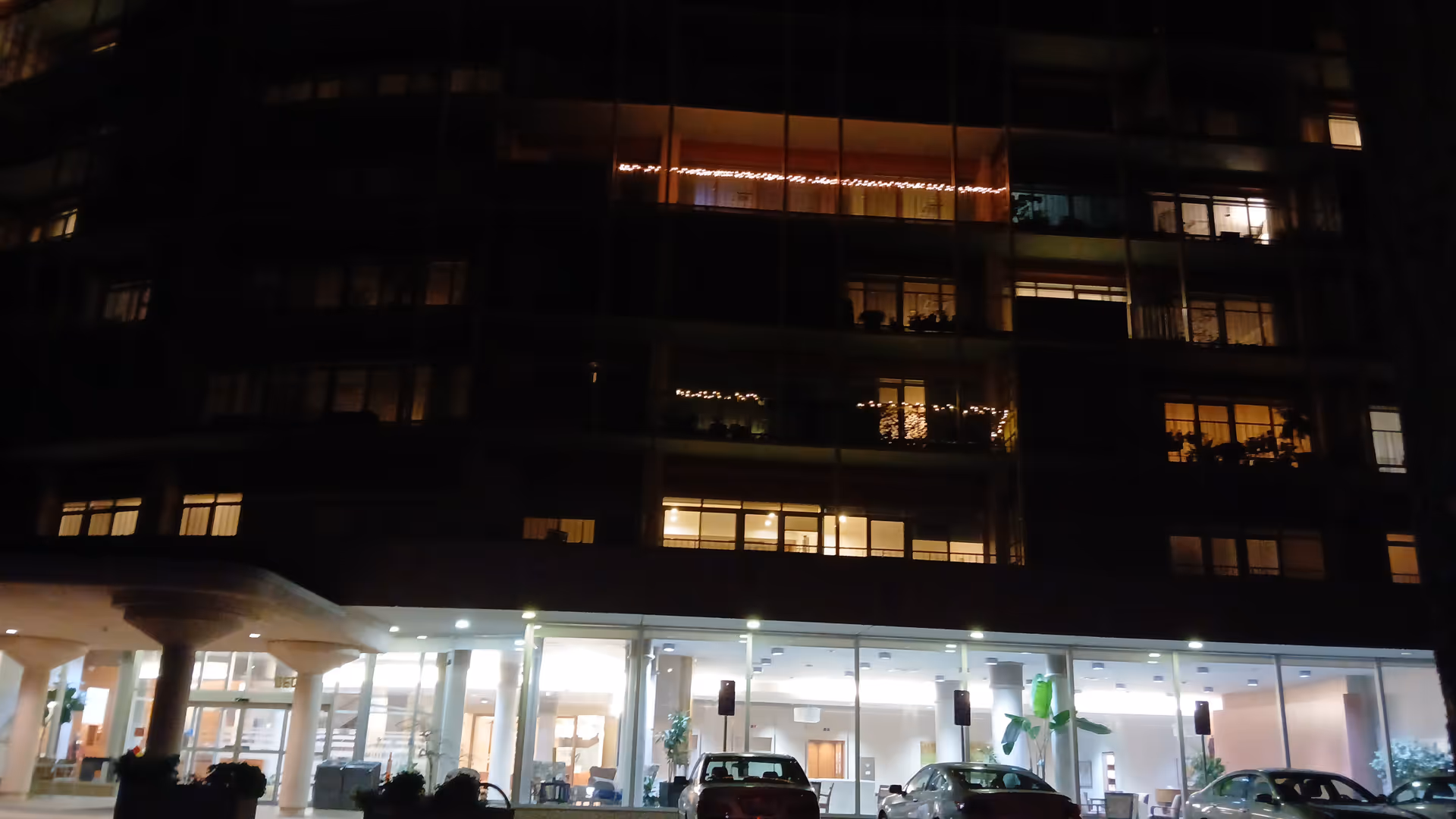 Nighttime exterior of a modern senior living facility with glass entrance and parked cars.