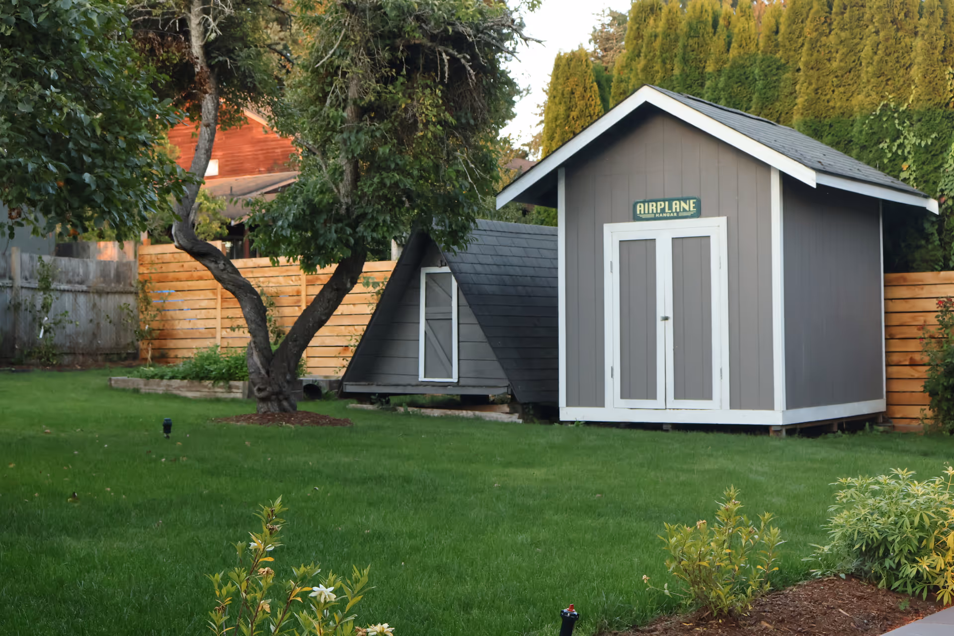 Green lawn with a gray shed and small outbuildings in a fenced backyard.