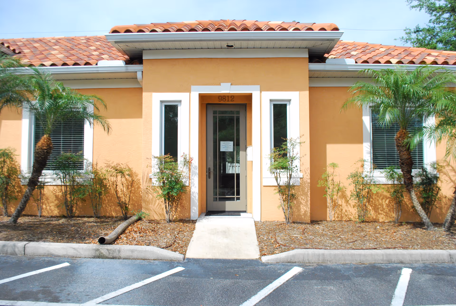 Orange stucco building entrance with white trim, small shrubs, and palm trees outside.