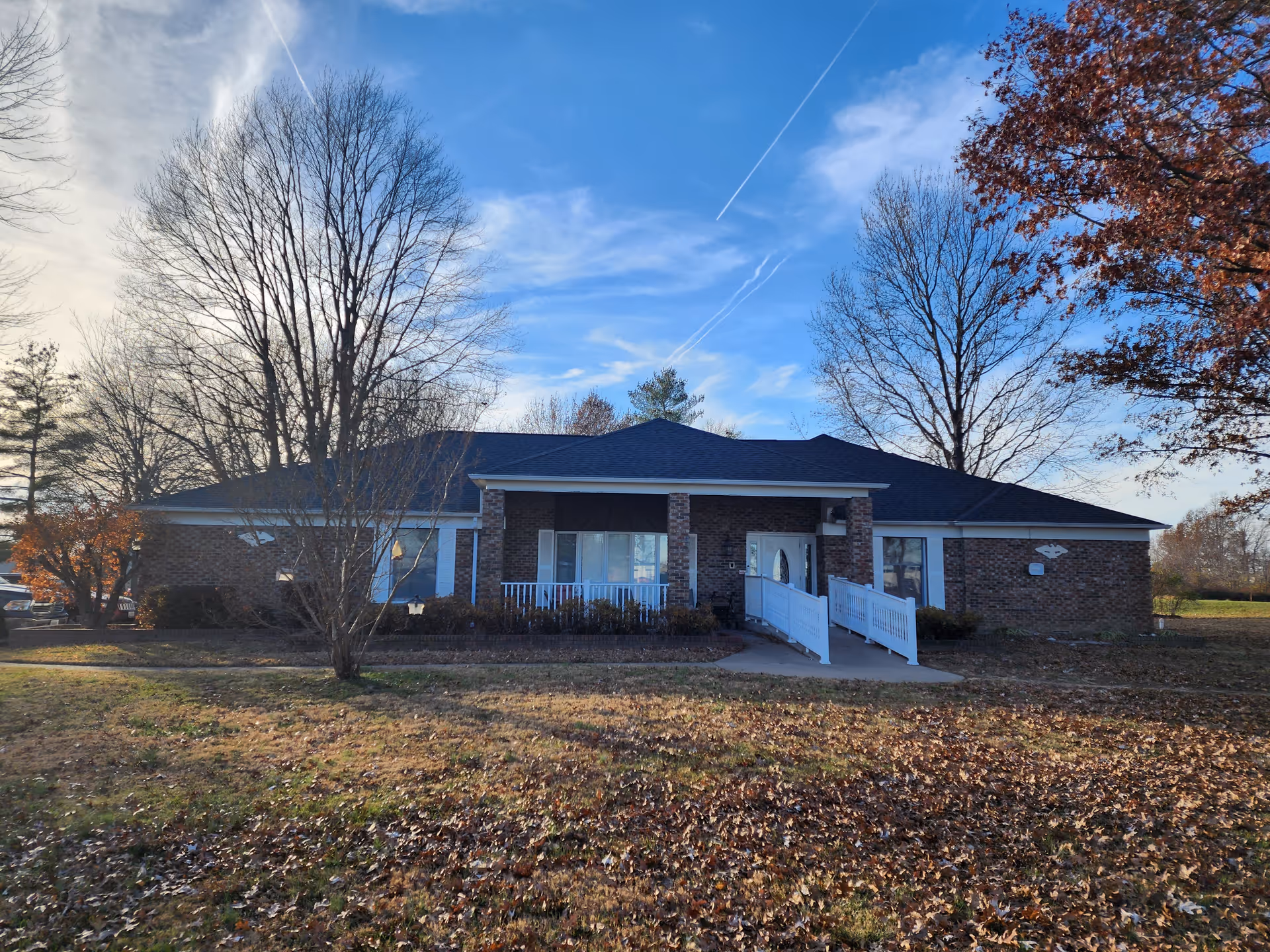 Single-story brick building with a front ramp and white railing surrounded by leaf-covered grass and bare trees under a blue sky.