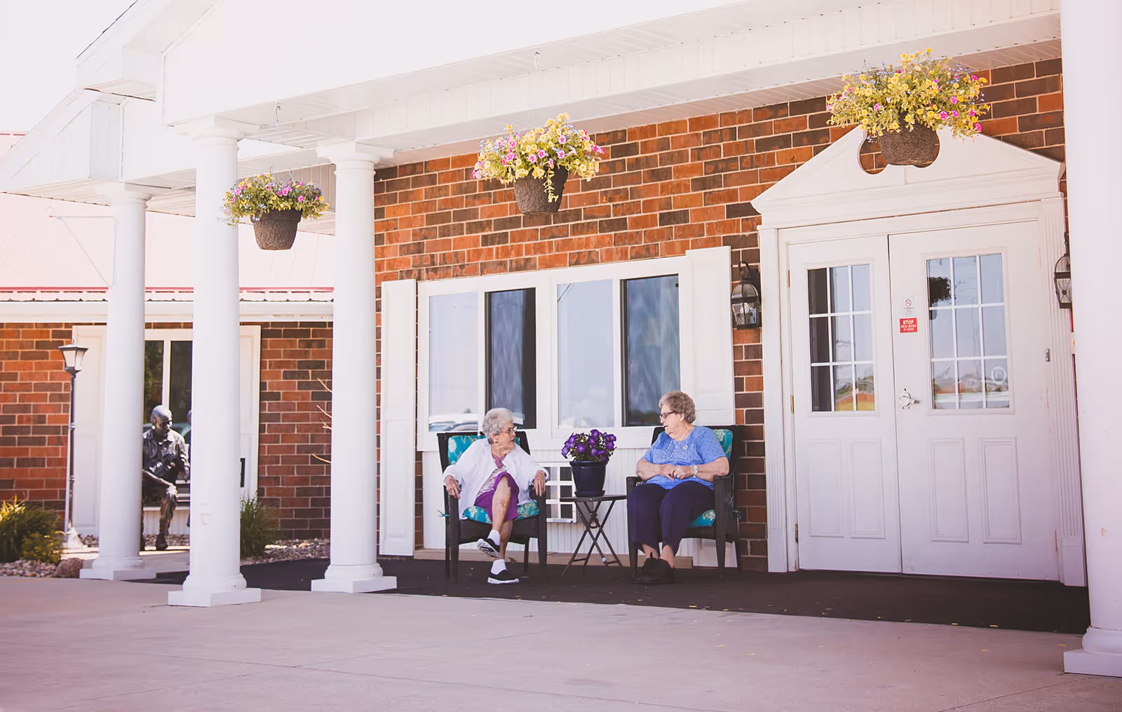 Two elderly women sitting and talking on chairs outside a brick building entrance with white columns and hanging flower baskets.