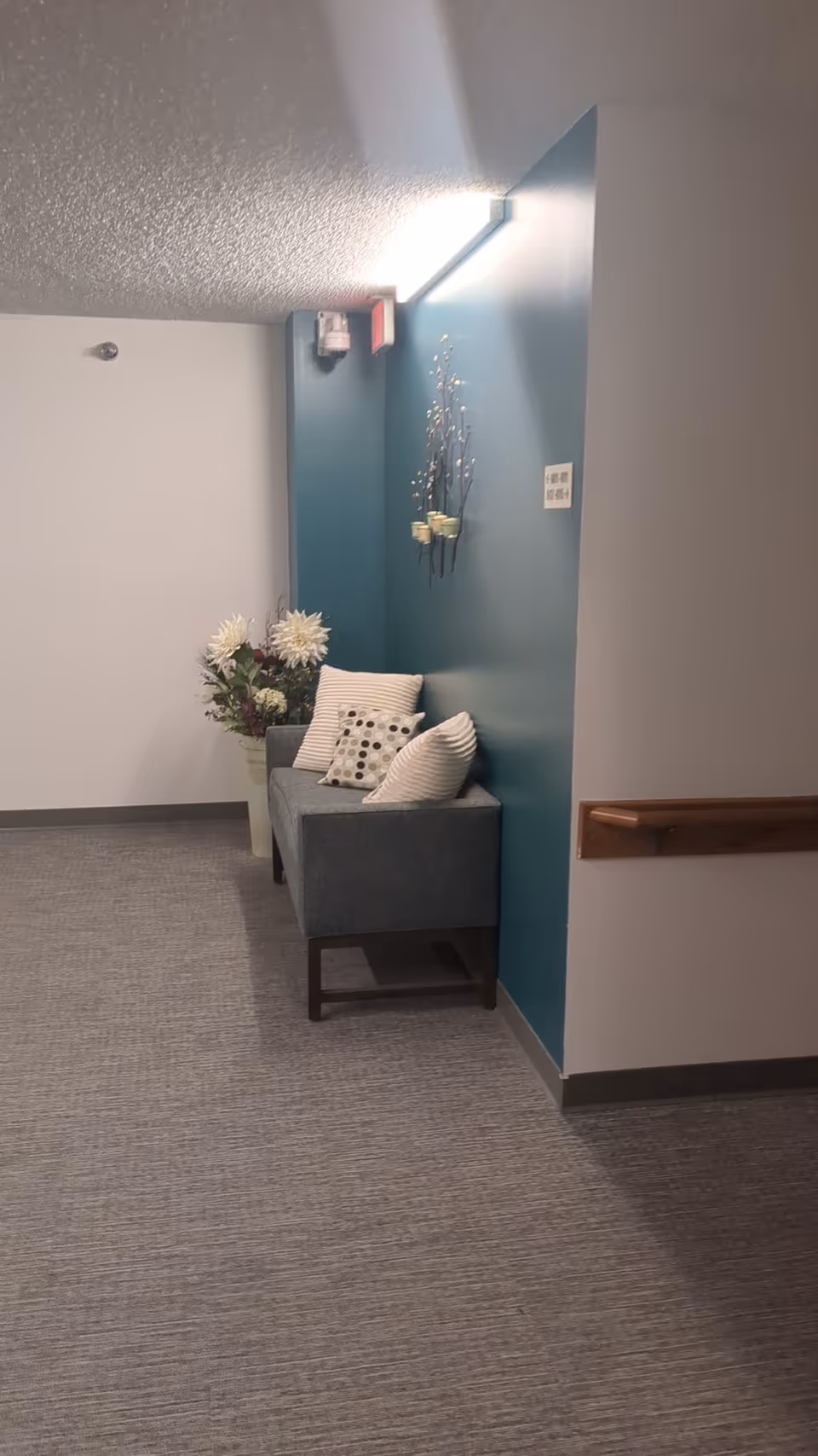 A hallway seating nook with a blue wall, chair, cushions, flowers, in a senior living facility