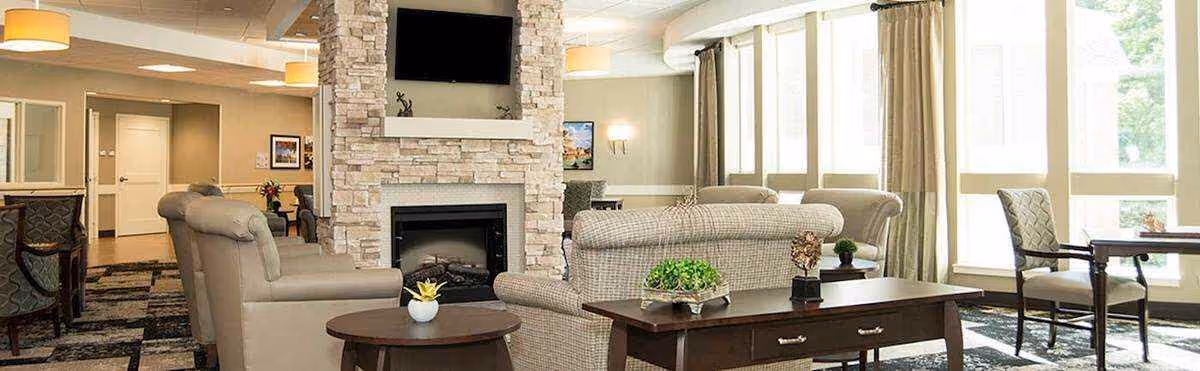 Bright common lounge with sofas and armchairs arranged around a stone fireplace and TV, with large windows and side tables.