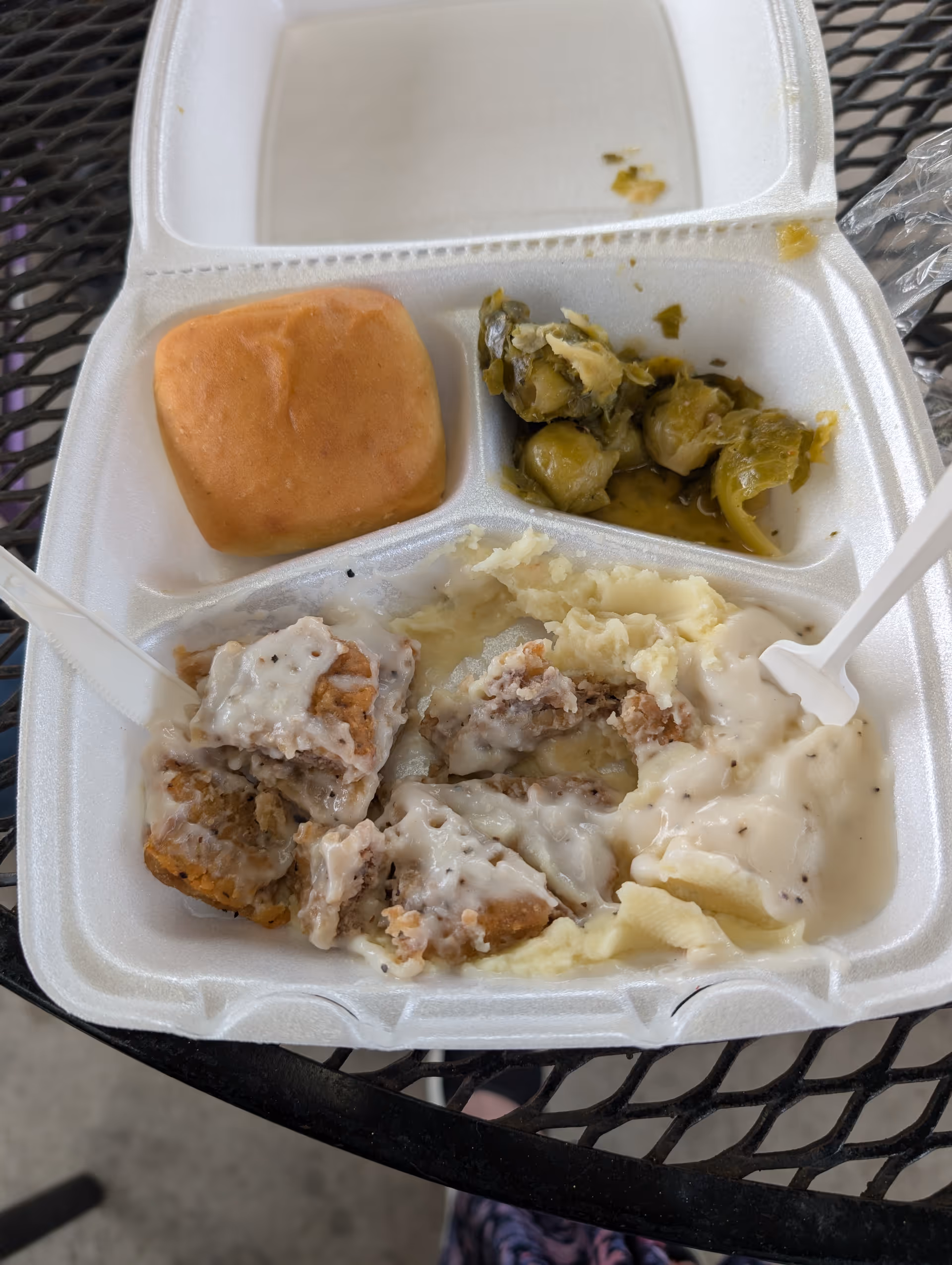 Styrofoam meal tray on outdoor table with mashed potatoes, meat, roll, and vegetables.