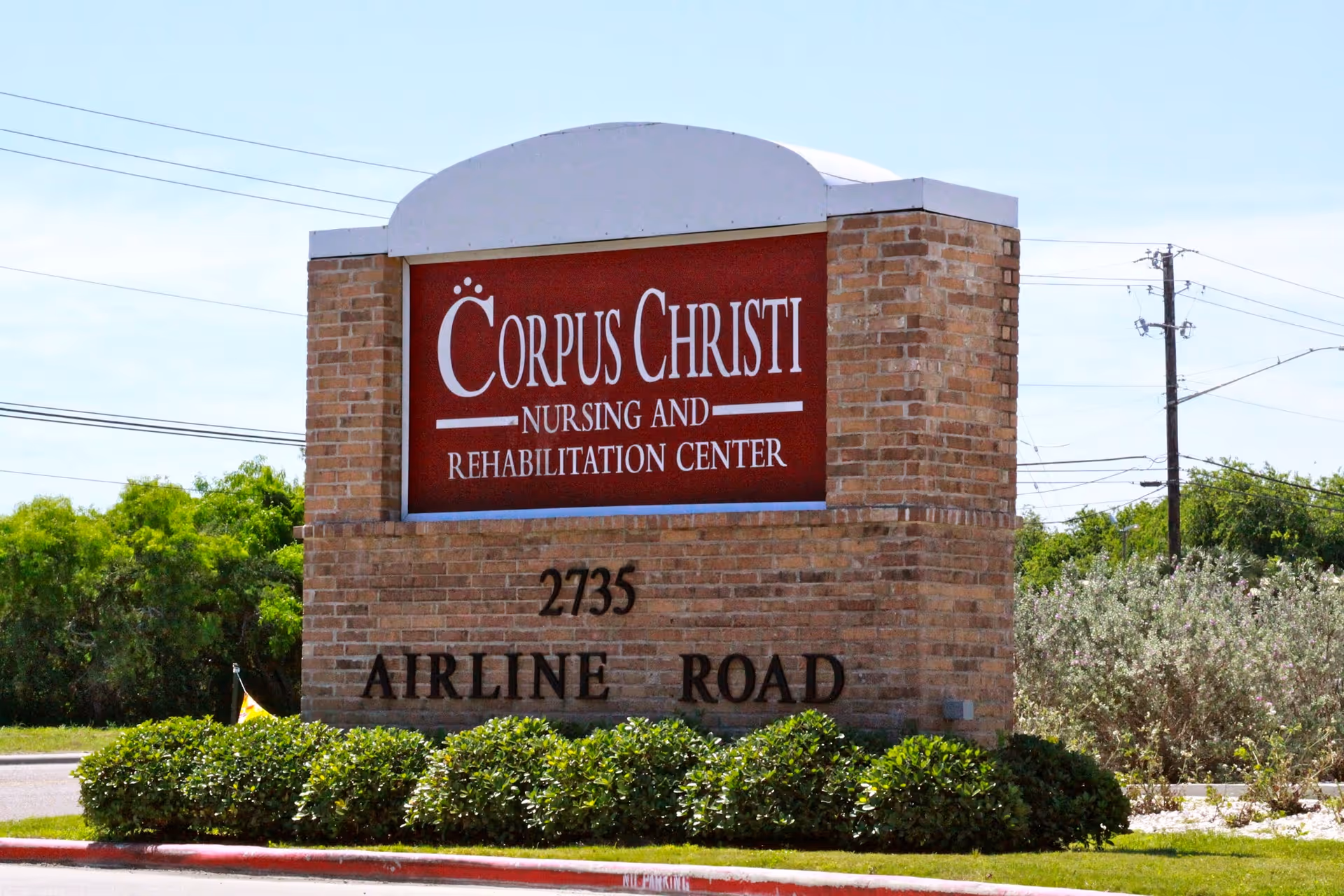 A brick sign for Corpus Christi Nursing and Rehabilitation Center located at 2735 Airline Road, surrounded by green bushes and trees under a clear sky.