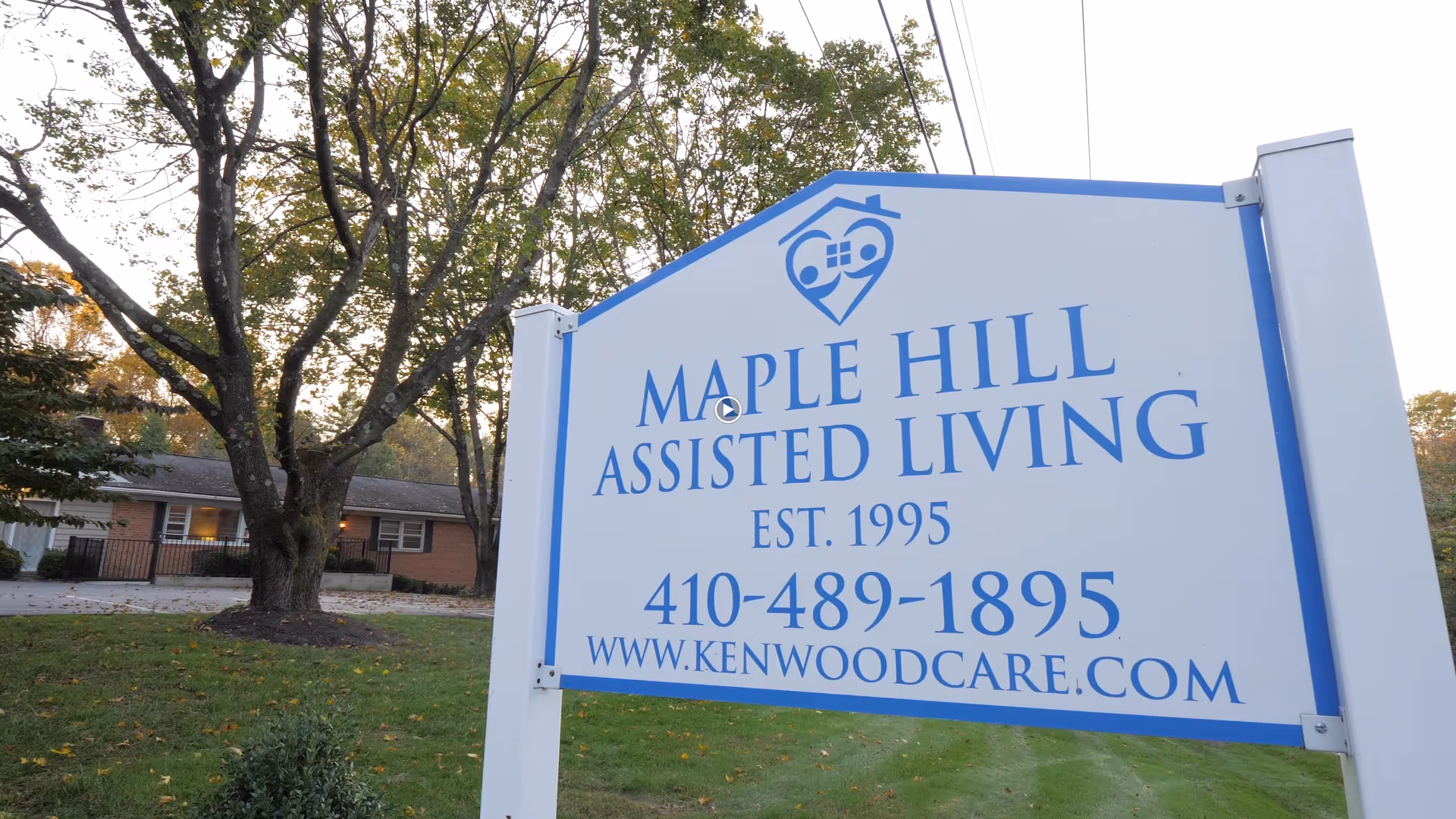 A large white and blue sign for Maple Hill Assisted Living with contact information and website, set on a grassy area with trees and a building in the background.