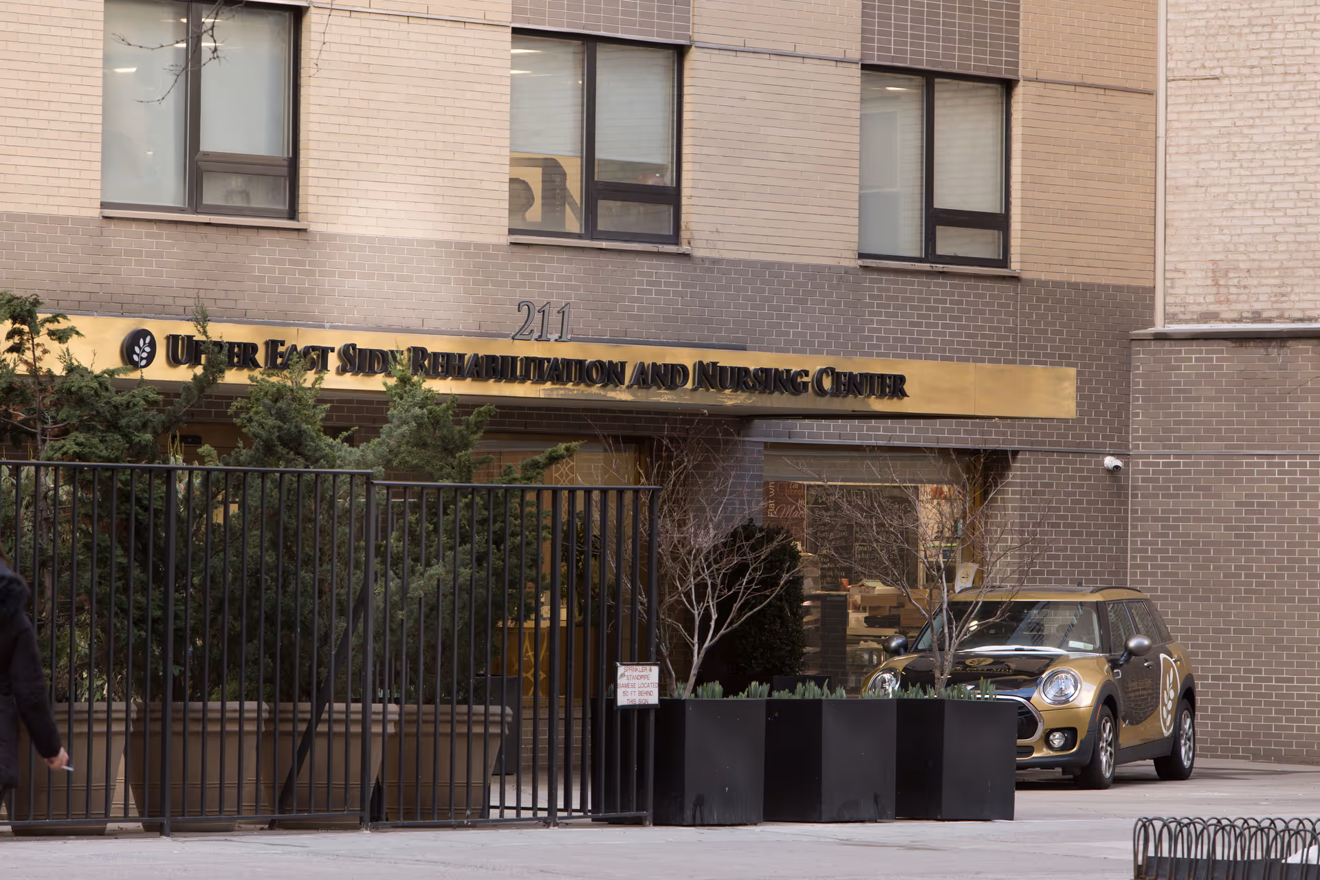 Exterior view of the Upper East Side Rehabilitation and Nursing Center building entrance with a gold sign displaying the facility name above the doorway. There are several green bushes behind a black metal fence and a parked Mini Cooper car to the right.