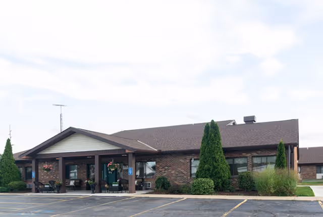 Exterior view of a single-story brick building with a covered entrance supported by columns. There are shrubs and small trees planted around the building, and an empty parking lot is visible in the foreground under a partly cloudy sky.