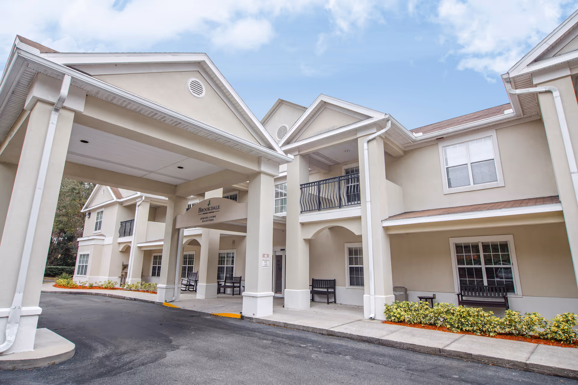 Exterior view of a beige senior living facility with a covered entrance and benches.