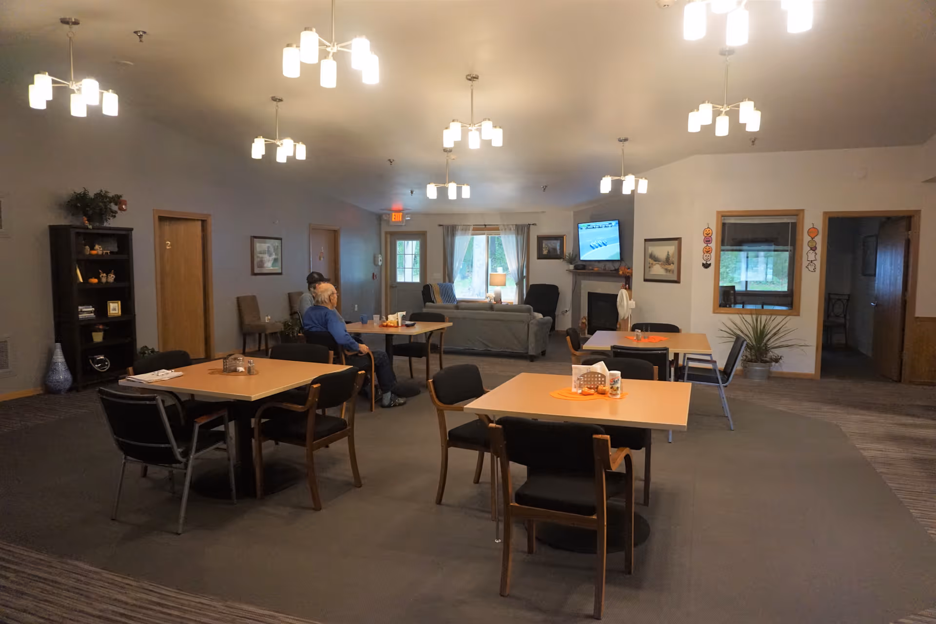 Interior senior living common area with tables, chairs, and a wall-mounted TV.