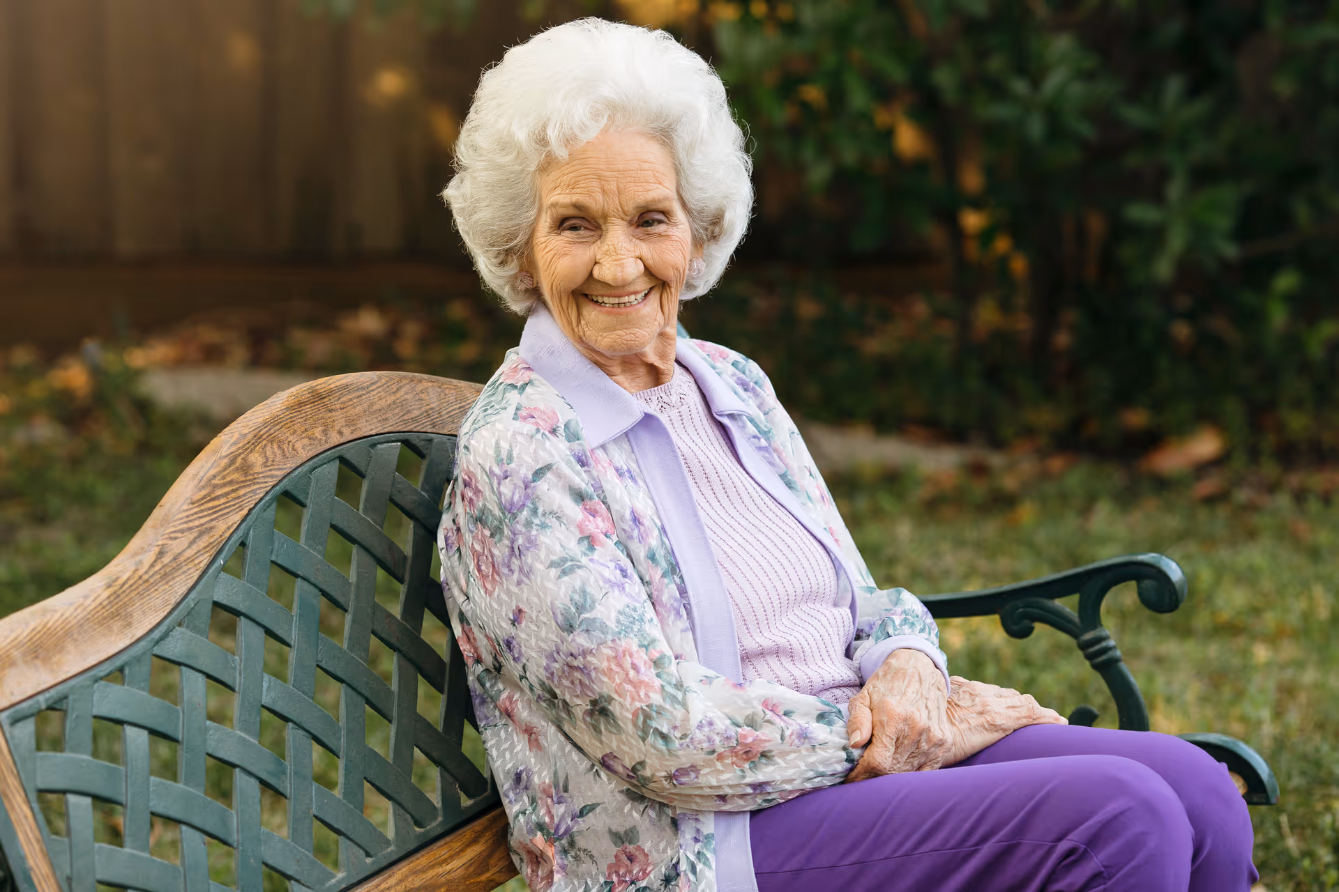 Smiling elderly woman sits on a wooden bench in a garden outdoors.