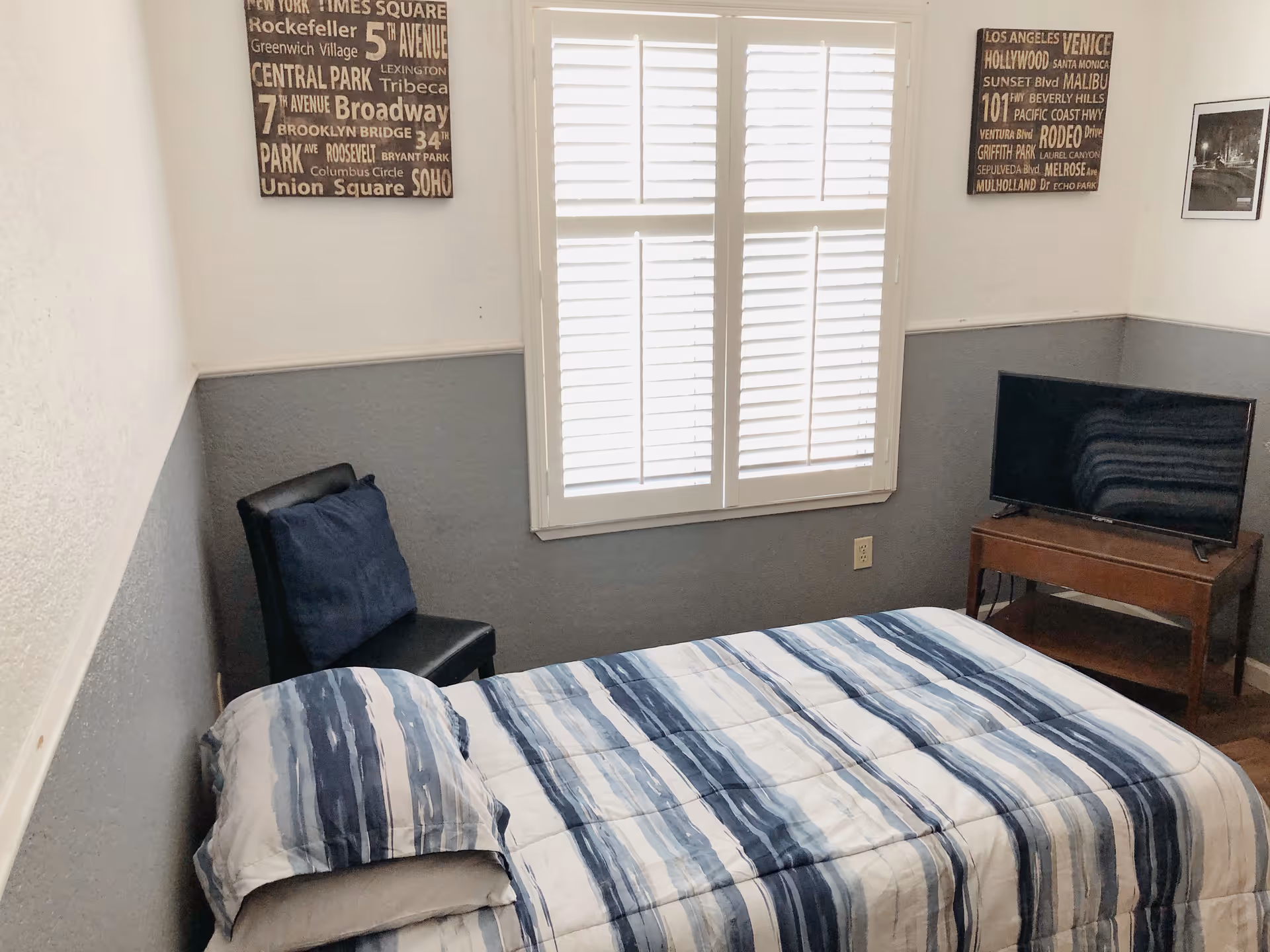 A compact bedroom with a blue-and-white striped bedspread, a small chair, a window with shutters, wall art, and a TV on a wooden stand.