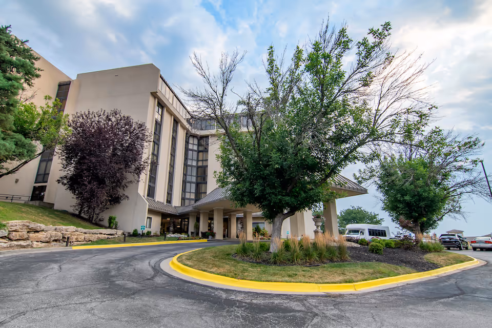 Front entrance of a multi-story senior living building with a covered drop-off, circular driveway, landscaping, and parked vehicles.