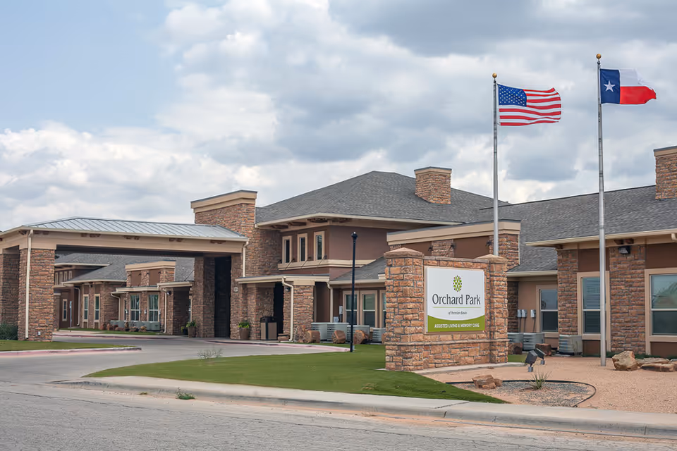 Front exterior of the Orchard Park assisted living facility showing the entrance portico, stone signage, and American and Texas flags.