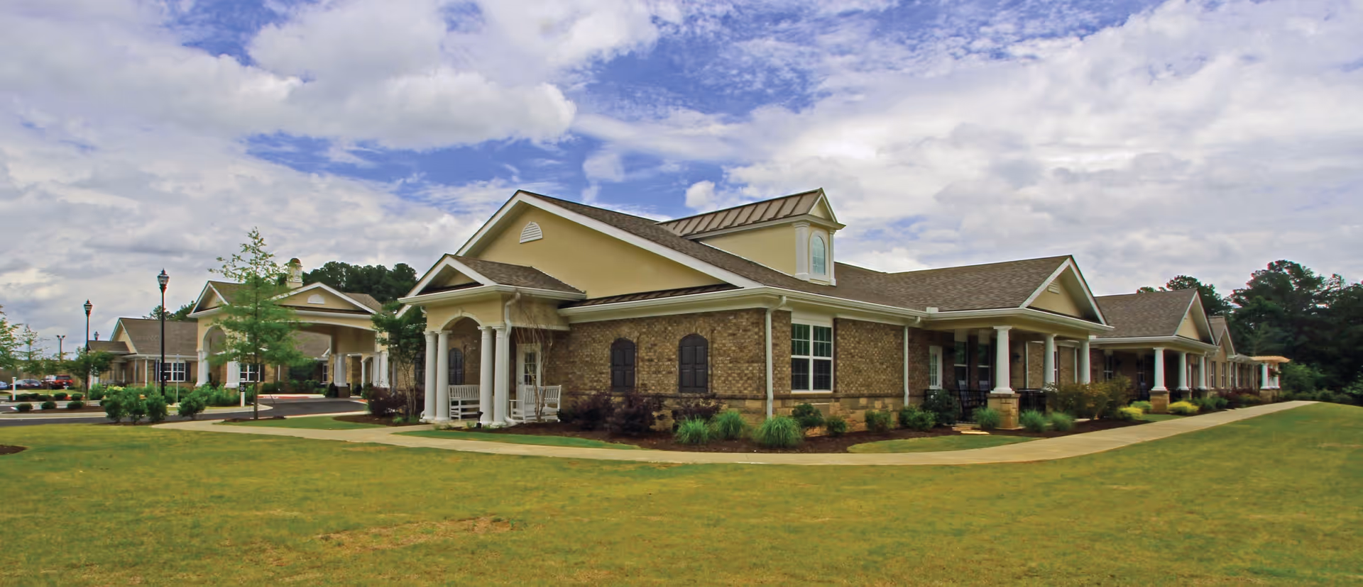 Single-story brick and stucco senior living building with columns, covered porches and a manicured lawn under a partly cloudy sky.