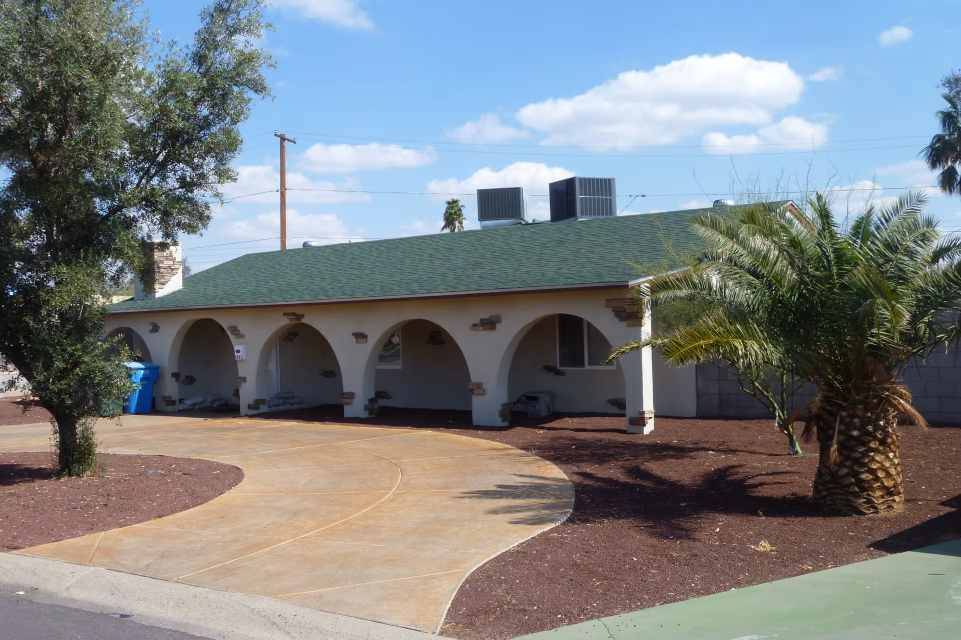 Single-story building with a green roof and arched front porch, surrounded by a landscaped yard with palm trees and a curved driveway under a partly cloudy sky.