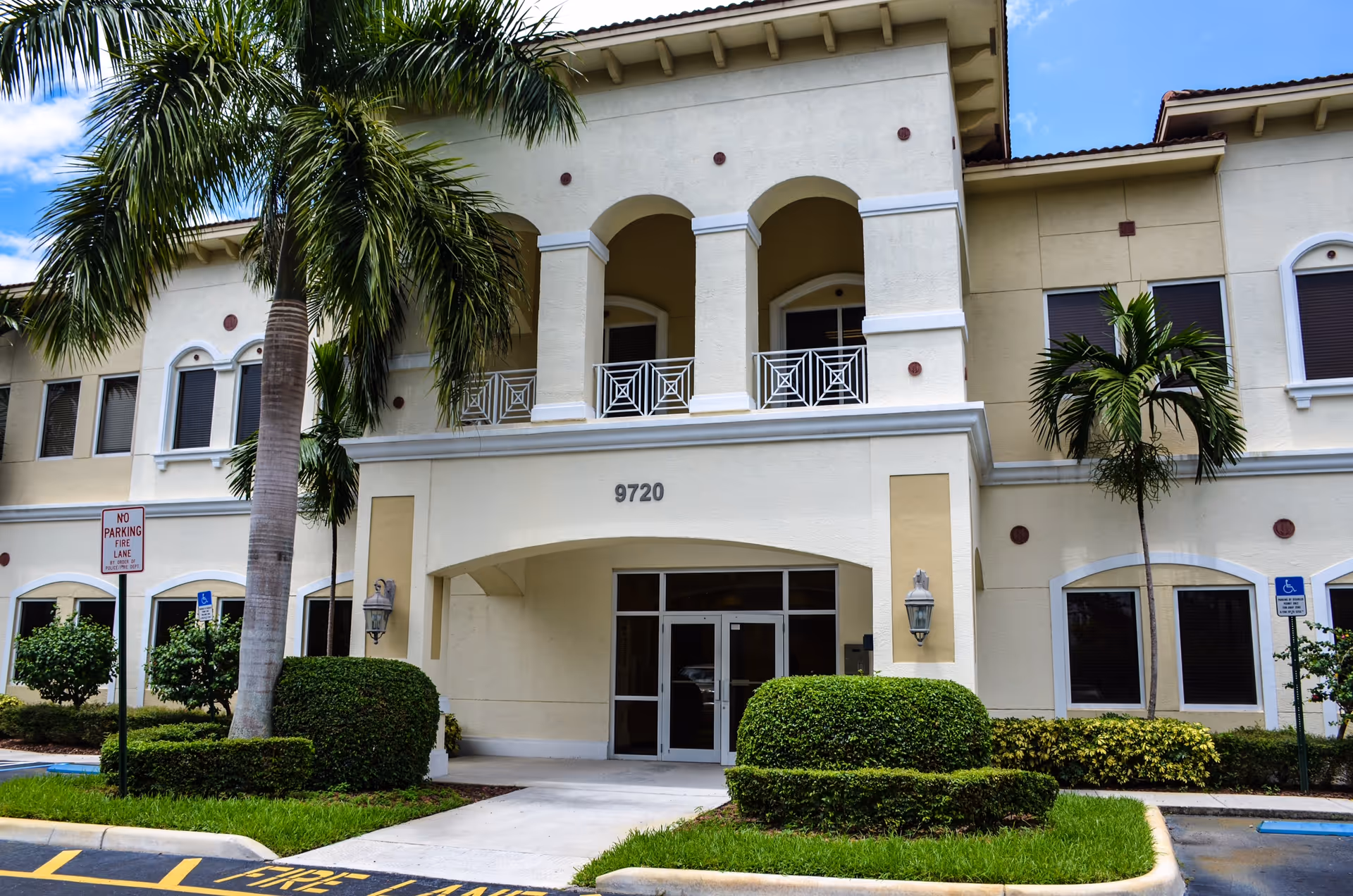 Front view of a beige senior living building with arches, palm trees, and landscaping.