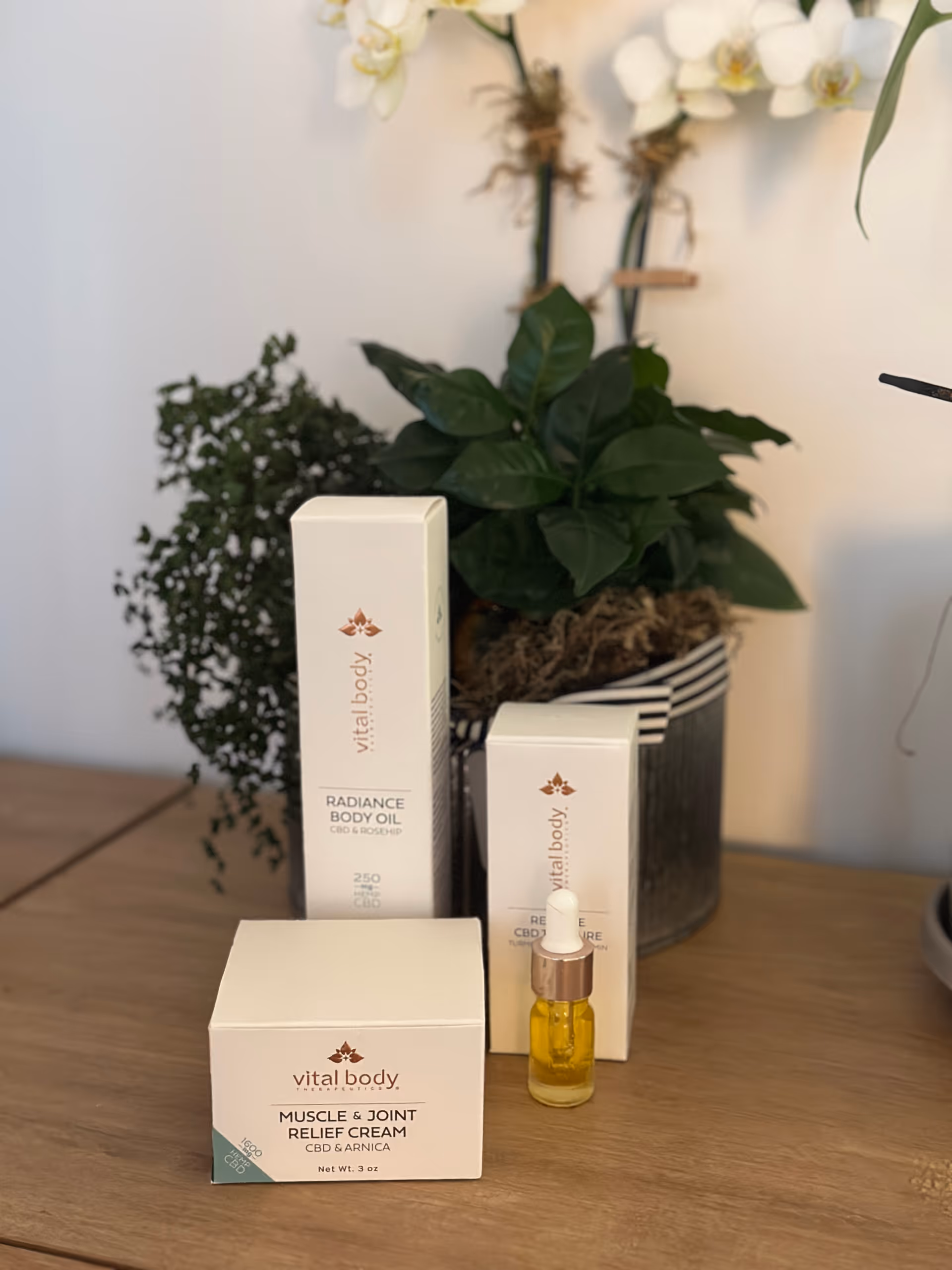 Arranged Vital Body skincare boxes and a small bottle on a wooden table with plants.