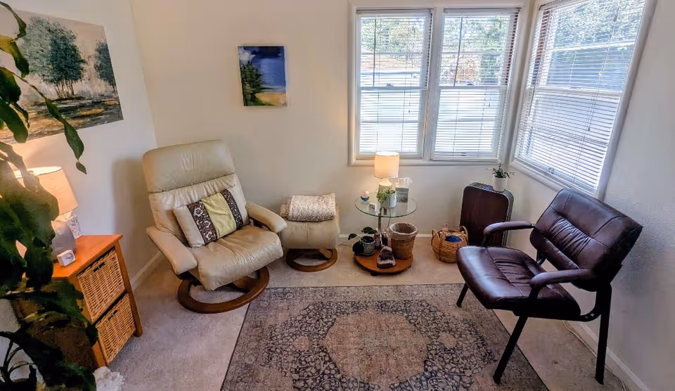 Cozy living room with beige recliner, brown chair, glass table, window blinds, and rug.