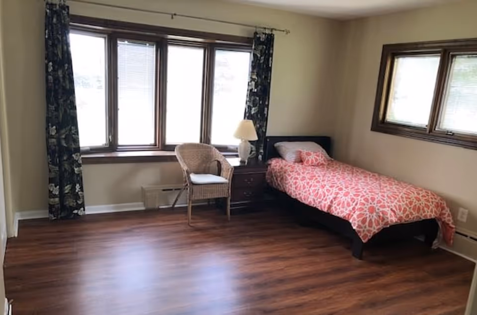Bright bedroom with a single bed, nightstand, chair, lamp, and two windows.