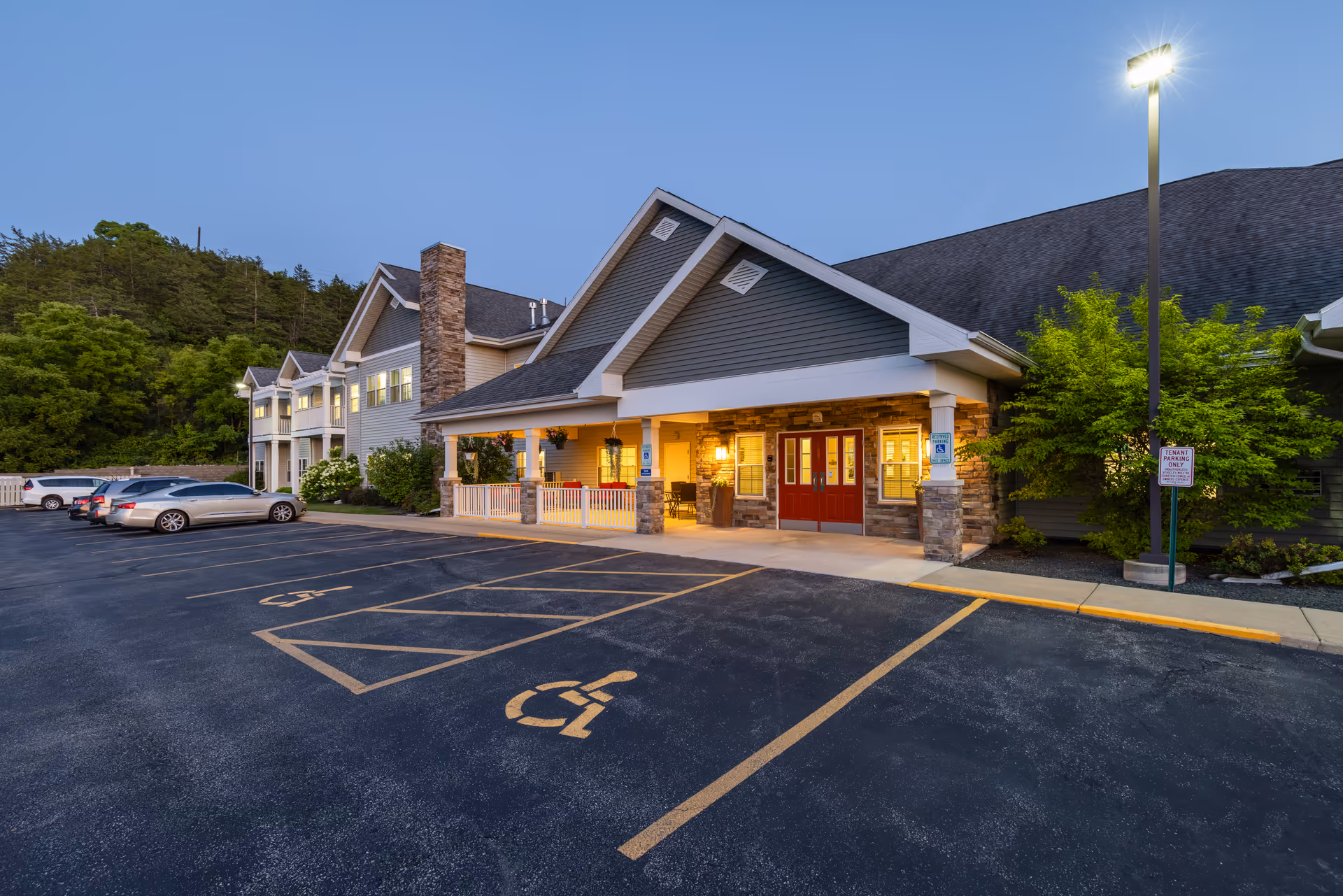 Exterior view of Milestone Senior Living Cross Plains building at dusk, showing the entrance with red double doors, a covered porch with stone pillars, several parked cars, and a well-lit parking lot with handicap parking spaces.