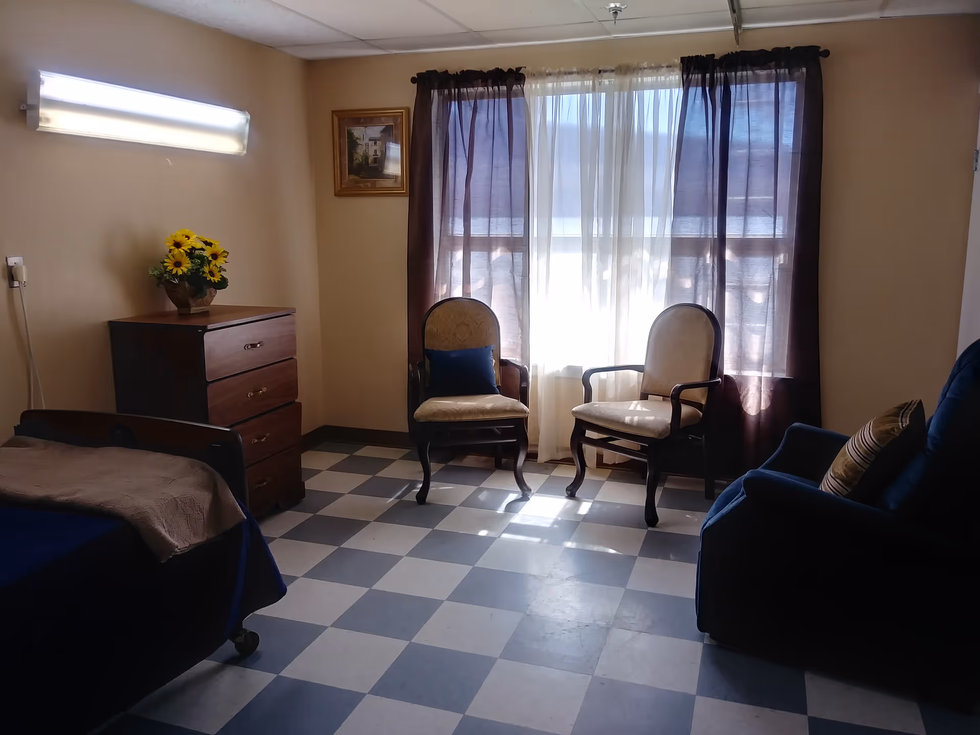 Sunlit resident bedroom with two upholstered chairs by a curtained window, a dresser topped with sunflowers, and a bed.