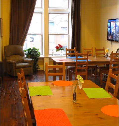 A dining area with wooden tables and chairs, colorful placemats, a small vase with a flower on the table, a large window with brown curtains, a potted plant, a comfortable armchair, and a wall-mounted television showing a news program.