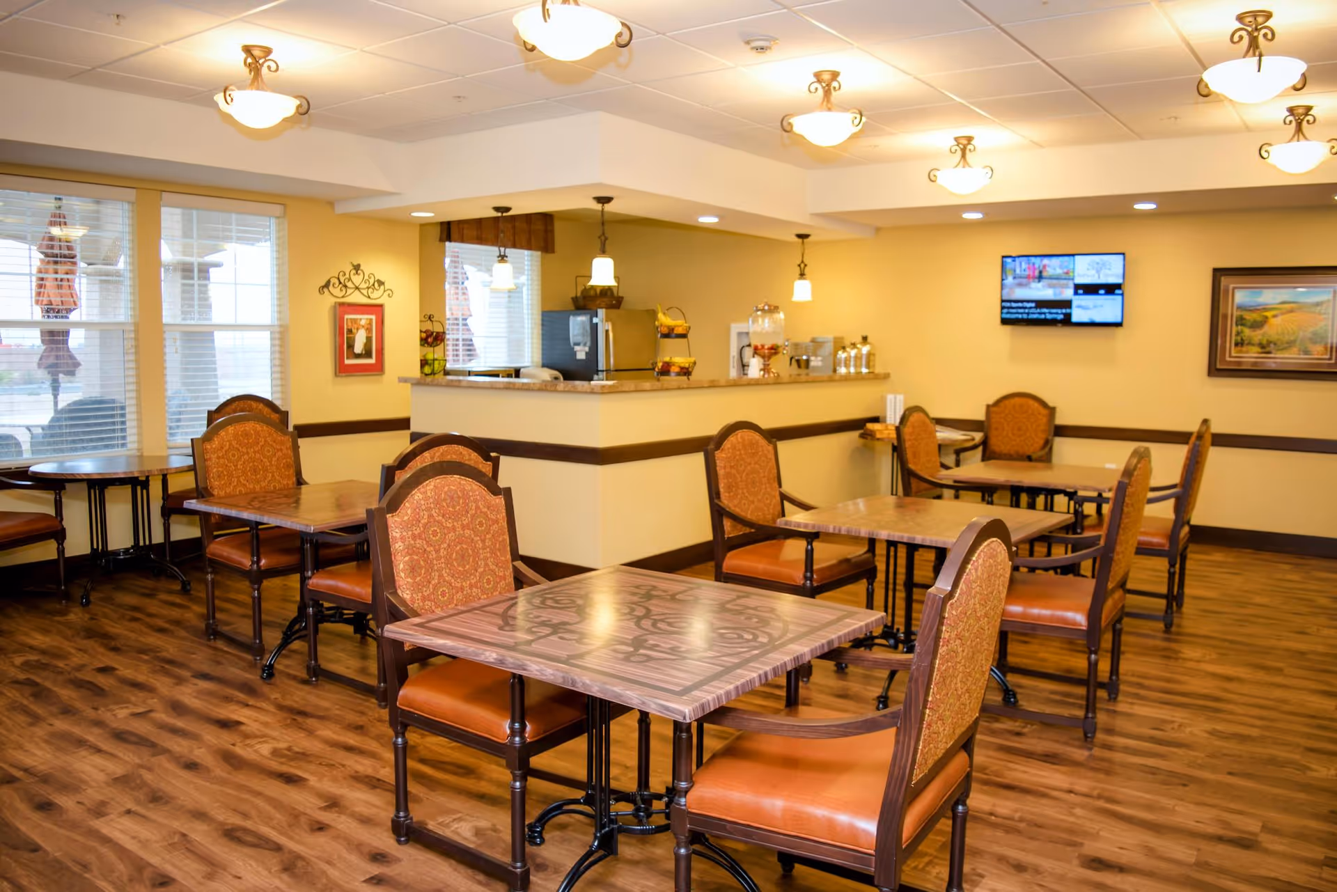 A well-lit dining area with several square tables and cushioned chairs with patterned upholstery. The room has wooden flooring, cream-colored walls, and multiple ceiling light fixtures. A counter area with a coffee machine and water dispenser is visible in the background, along with a wall-mounted TV and framed artwork.