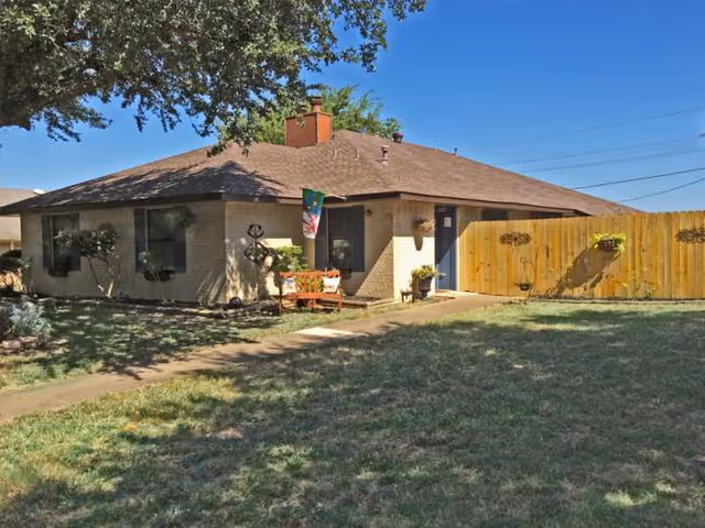 Single-story house exterior with lawn, wooden fence, and outdoor seating.